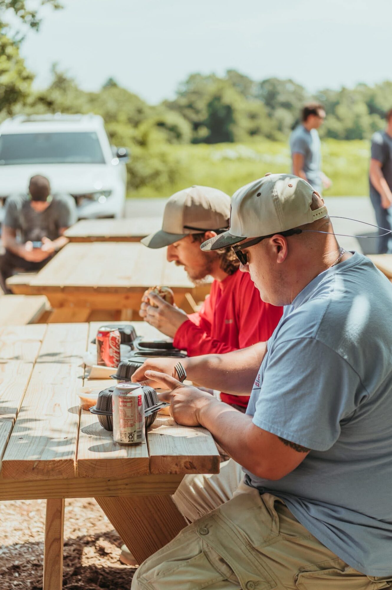 Three people sitting at a wooden table outdoors, using smartphones, with cans on the table, in a sunny setting.