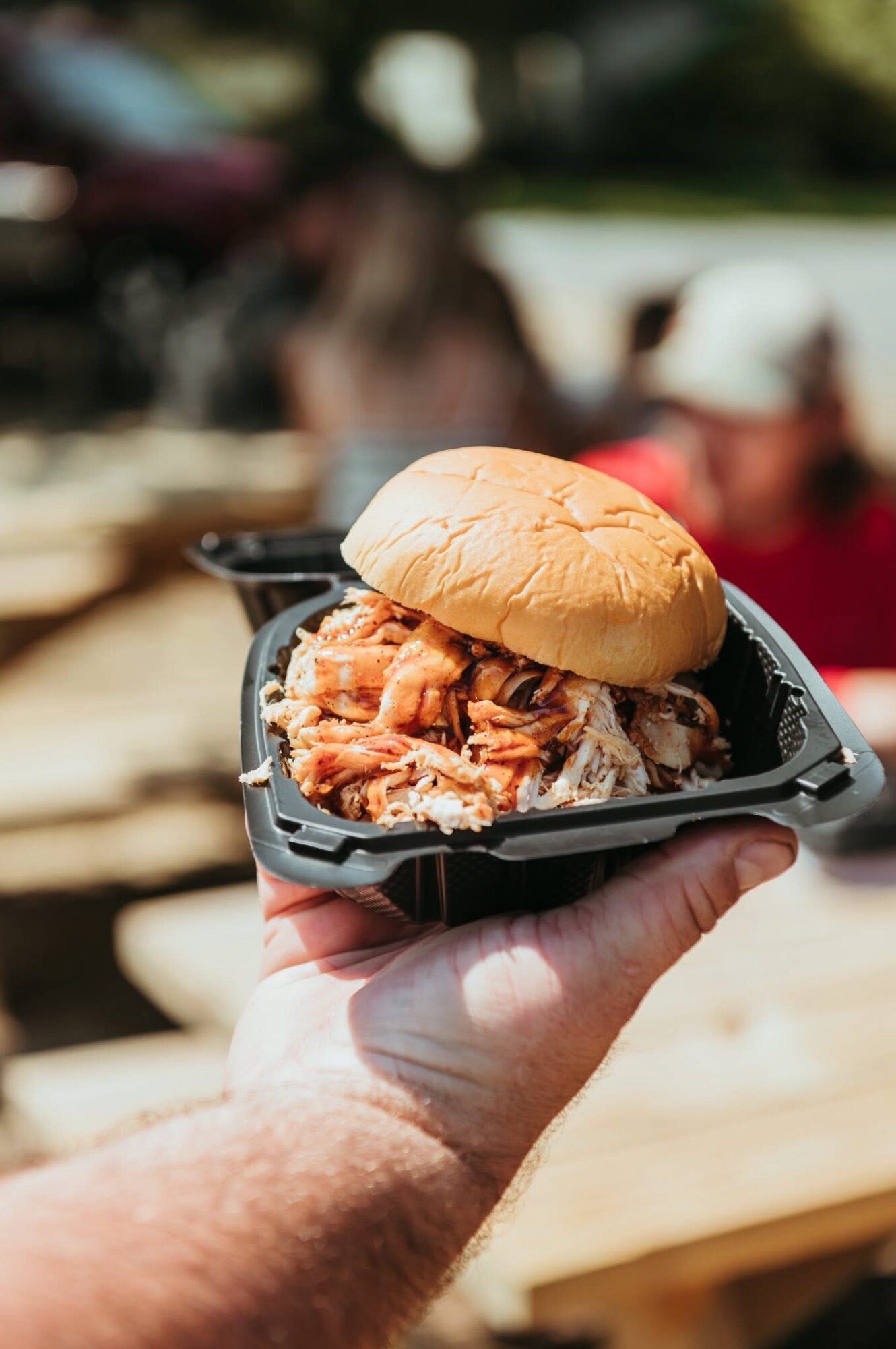 Hand holding a small black tray with a pulled pork sandwich in a bun outdoors, blurred background of people and trees