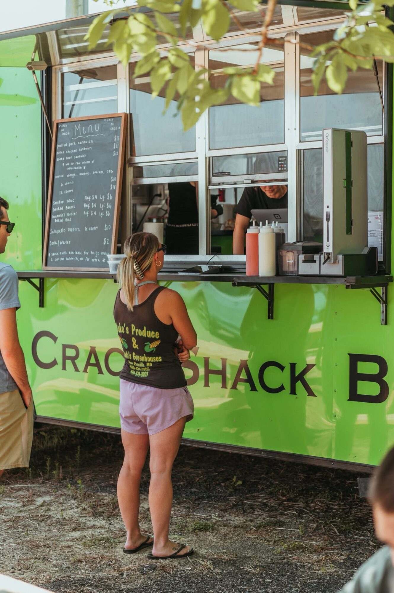 People order food at a green food truck with a menu board, window, and drinks, outdoors with trees and leaves visible.