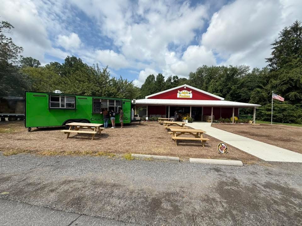 A red and white building with a porch, outdoor picnic tables, and a green trailer, under a partly cloudy sky.