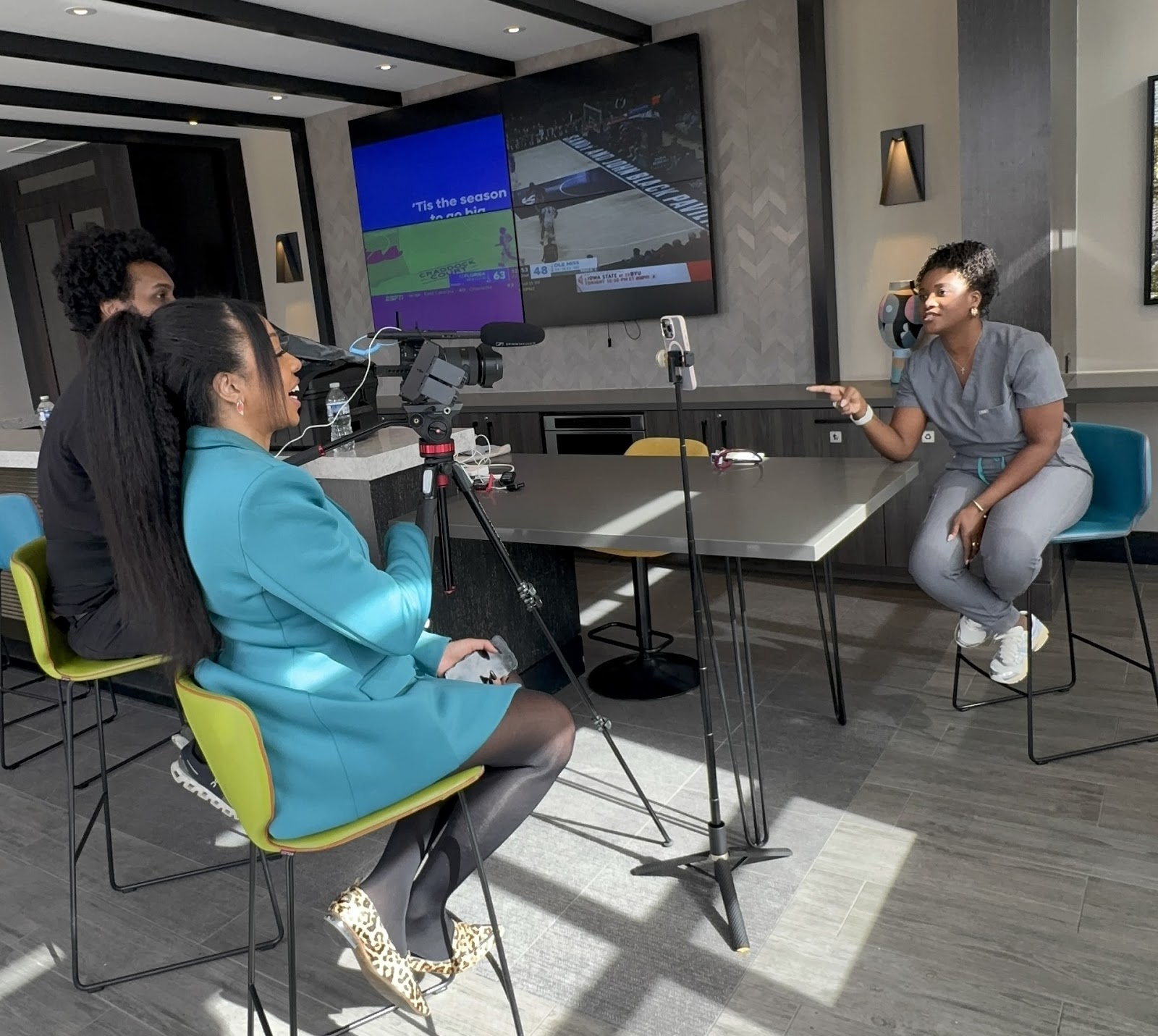 A woman in scrubs sits at a table, speaking to a camera crew with a large screen behind her, in a modern room.