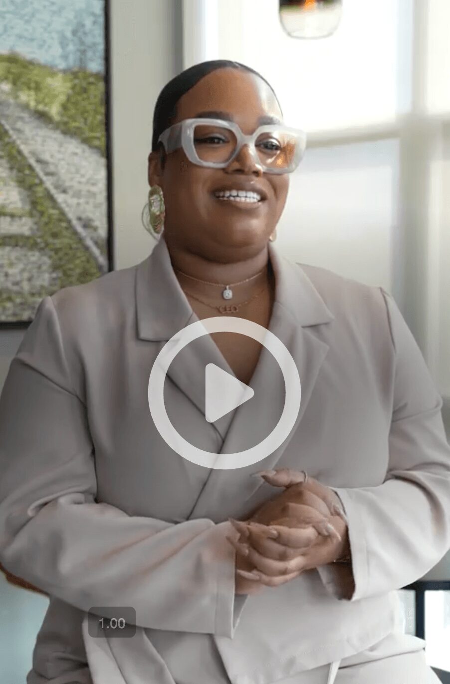 Woman with glasses and earrings smiling, wearing a beige blazer, in a bright room with a window and artwork behind her.