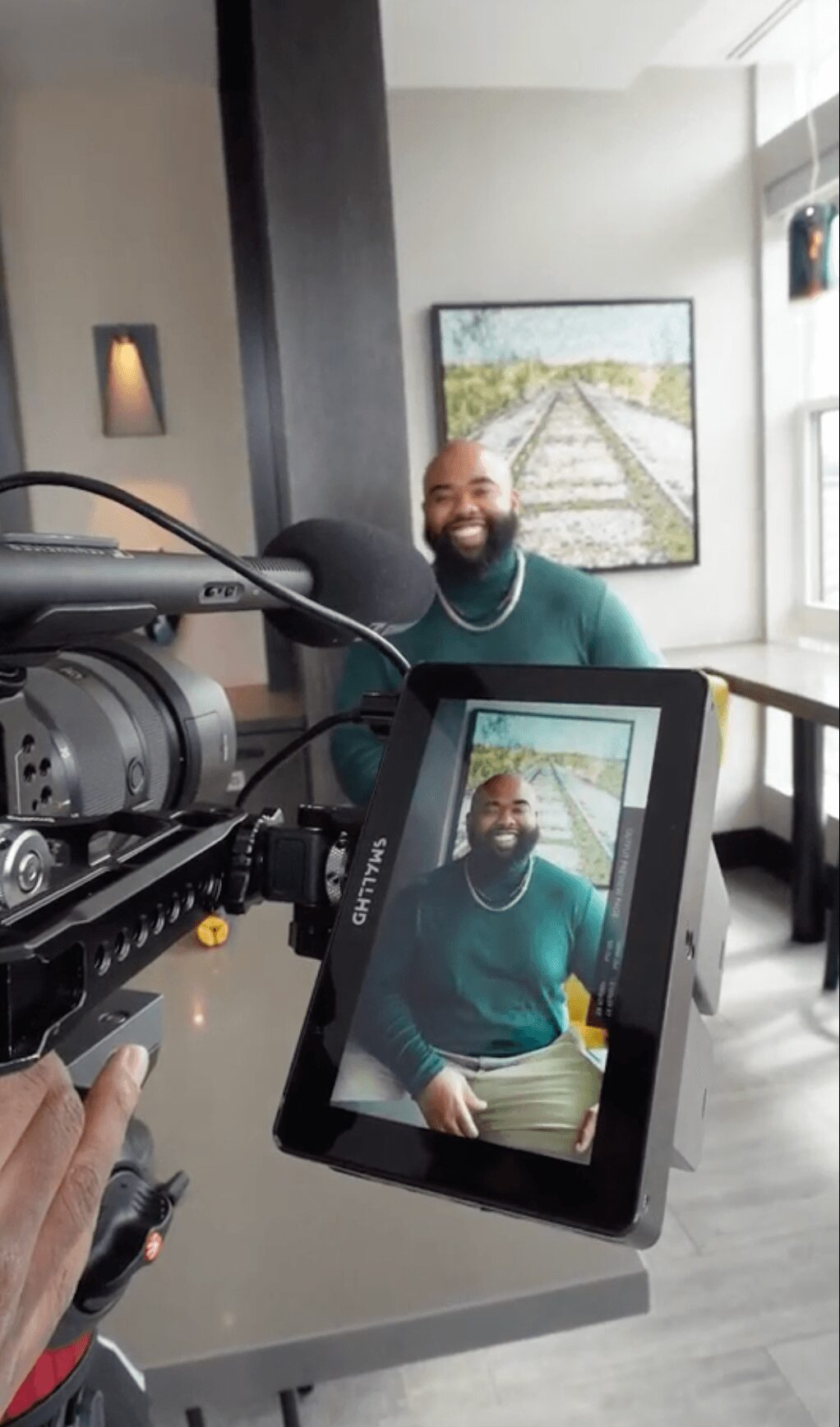 A man with a beard and green sweater is smiling, sitting indoors near a window with a painting behind him.