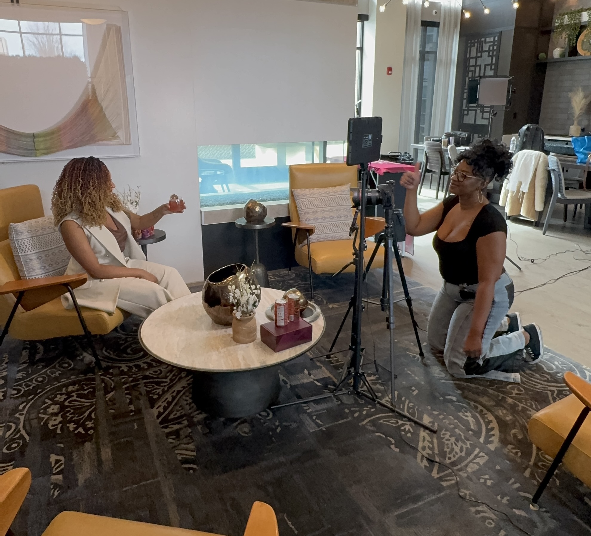 Two women in a room with chairs and a coffee table, one sitting and the other kneeling with a camera on a tripod, filming.