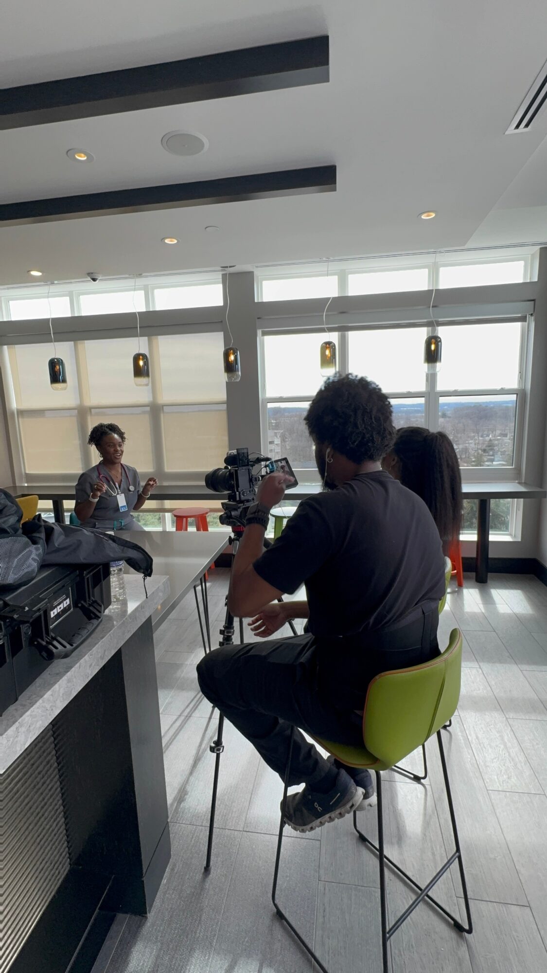 Person filming or photographing at a table in a bright room with large windows, other people seated nearby, and equipment on the table.