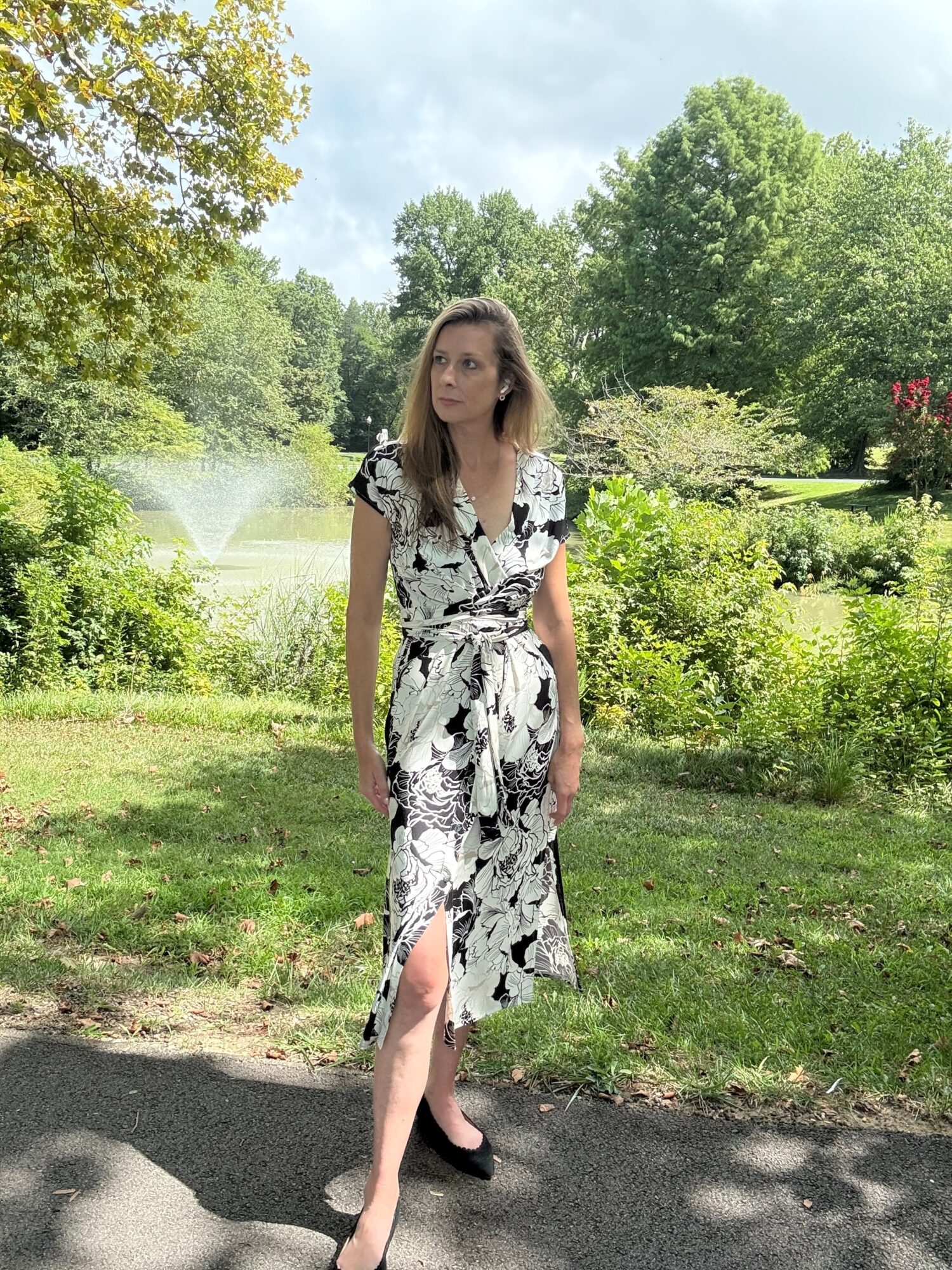 Woman in a floral dress standing outdoors in a park with trees and a pond in the background.