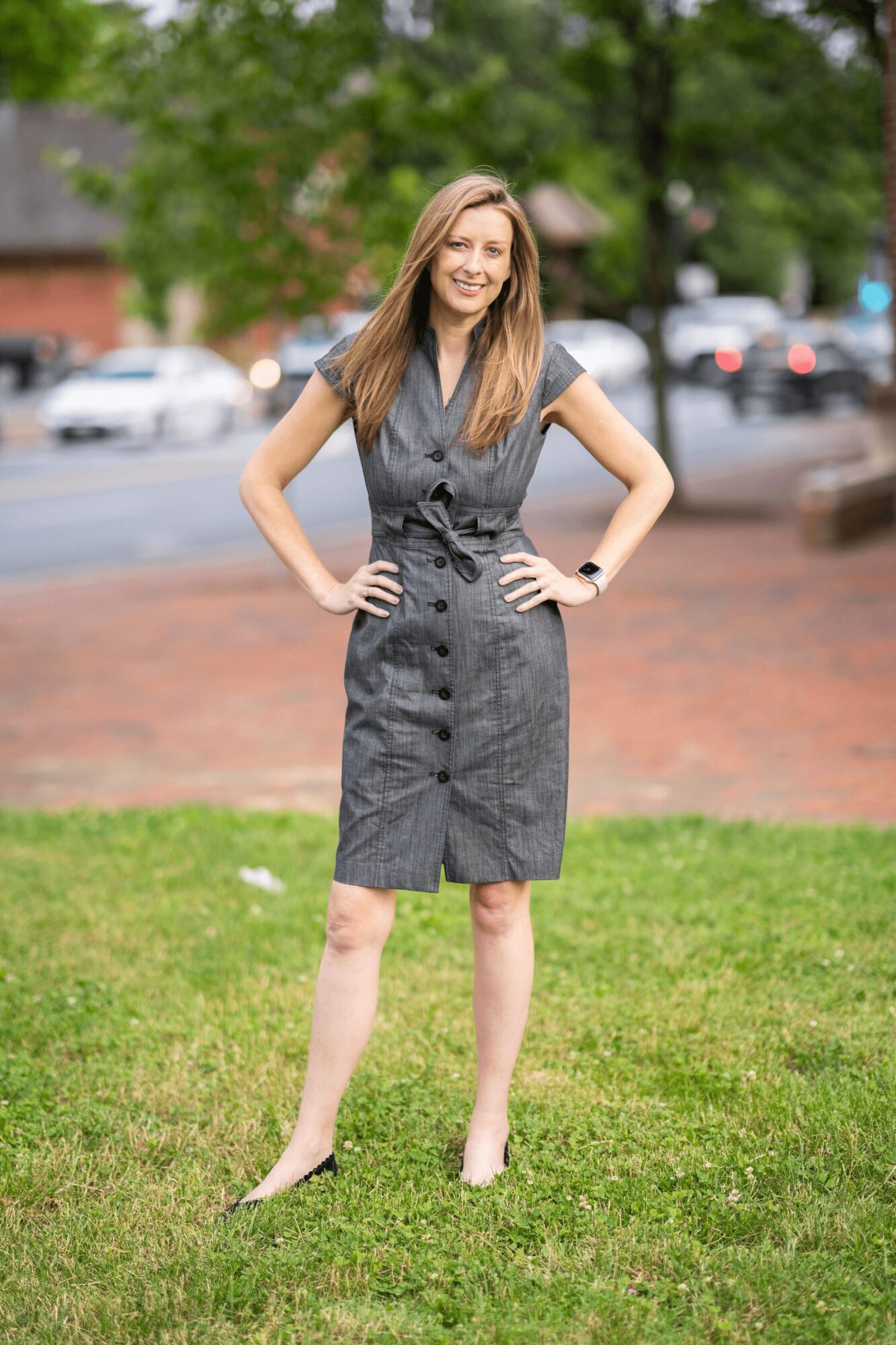 Woman standing outdoors on grass, smiling, hands on hips, wearing a gray dress, with trees and cars in background.