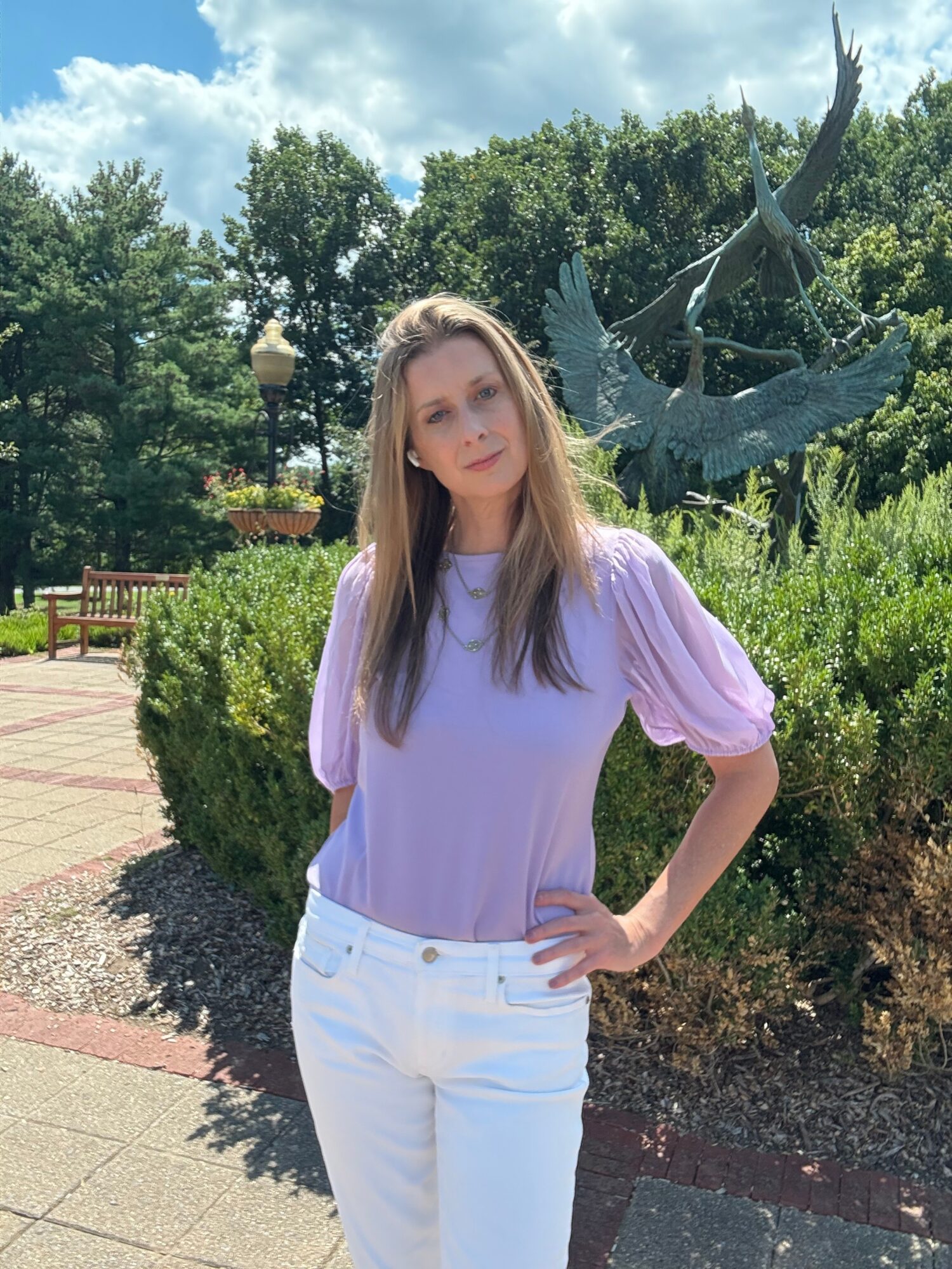 Young woman with long hair wearing a lavender blouse and white pants standing outdoors near a sculpture of birds, with trees and a park in the background.