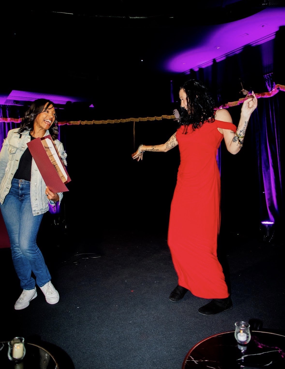Two women are on stage, one in a red dress holding a stick, the other with books, smiling at each other.