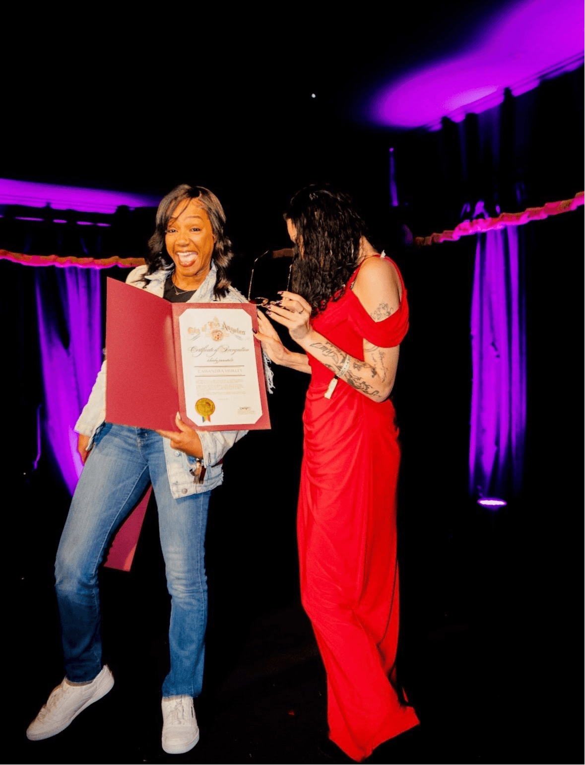 Two women on stage, one holding a certificate, smiling, with purple lighting in the background.