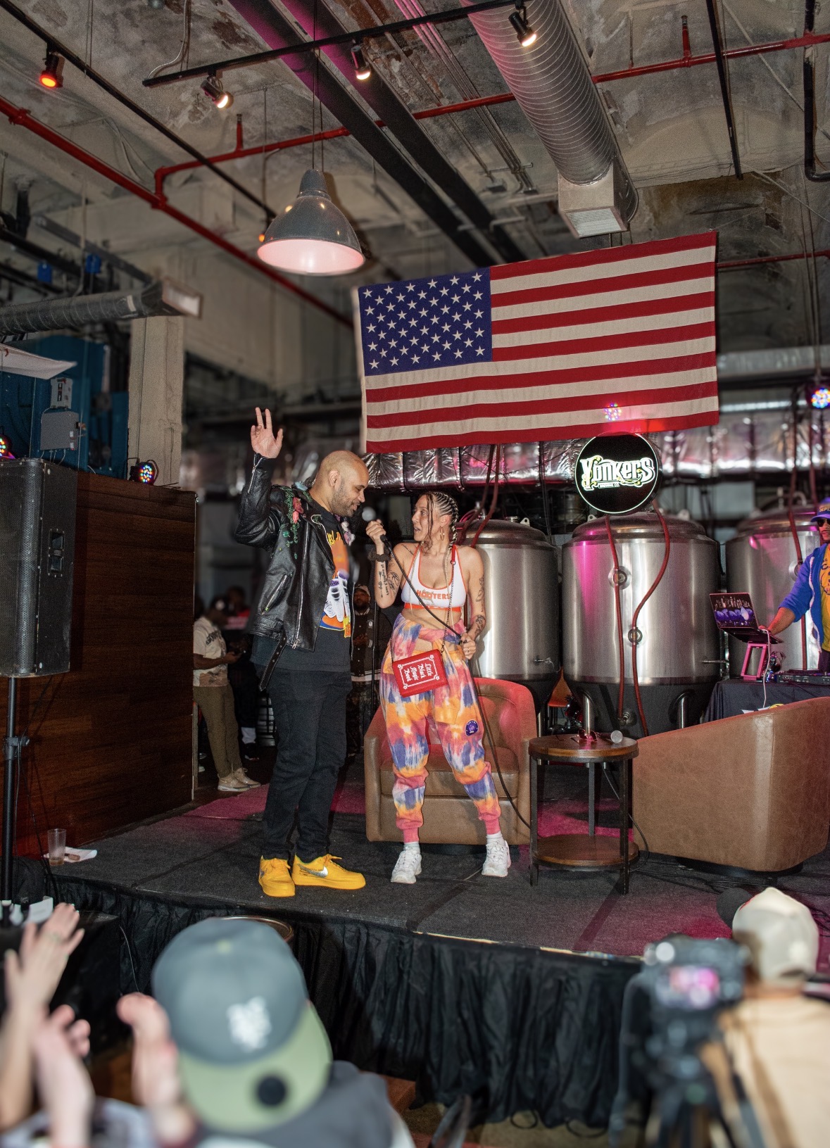 Two performers on stage with an American flag backdrop, one raising a hand, the other holding a microphone, audience in foreground.