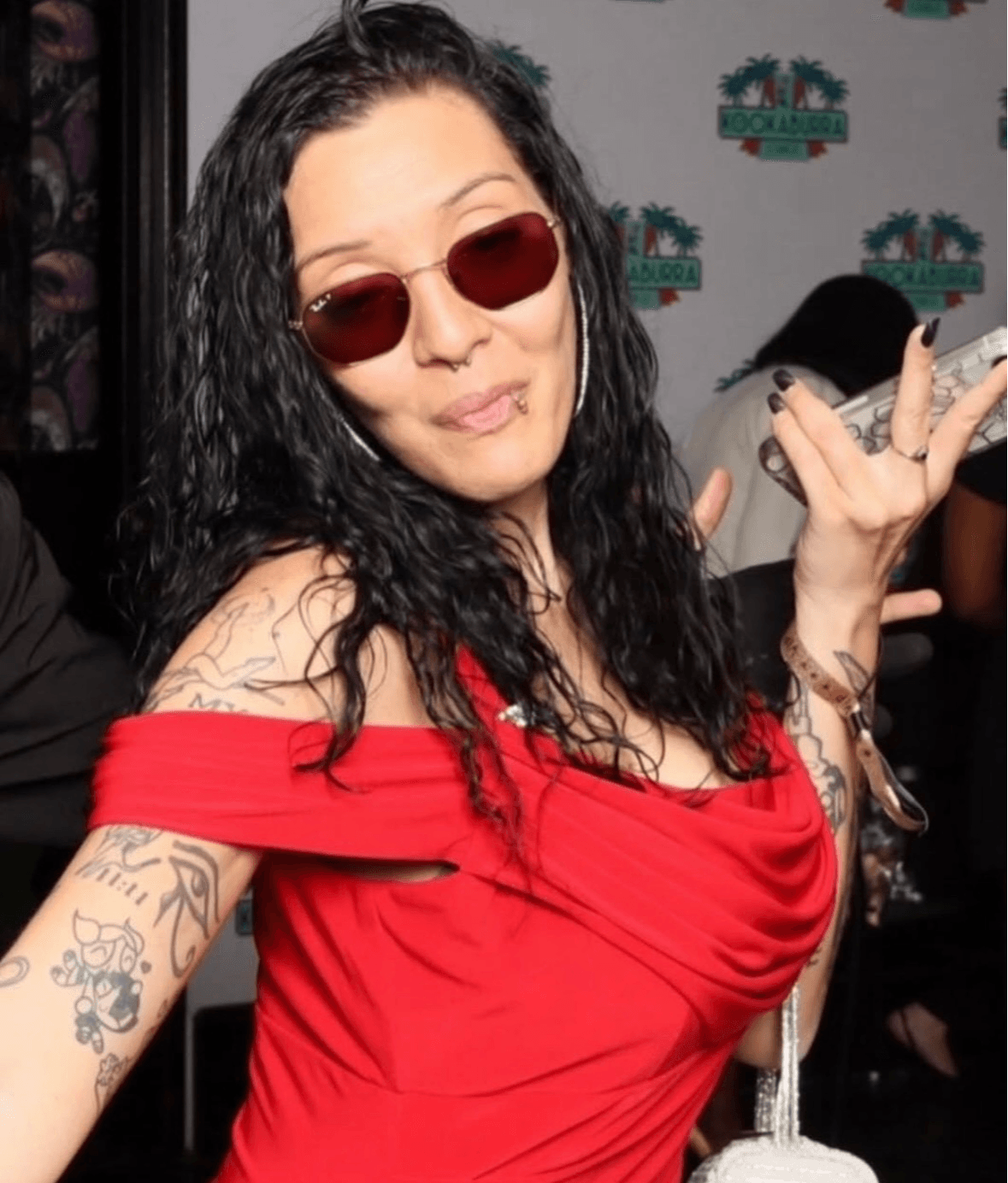 Woman with curly hair wearing sunglasses and a red off-shoulder top, posing with hand gestures, tattoos visible on arms.