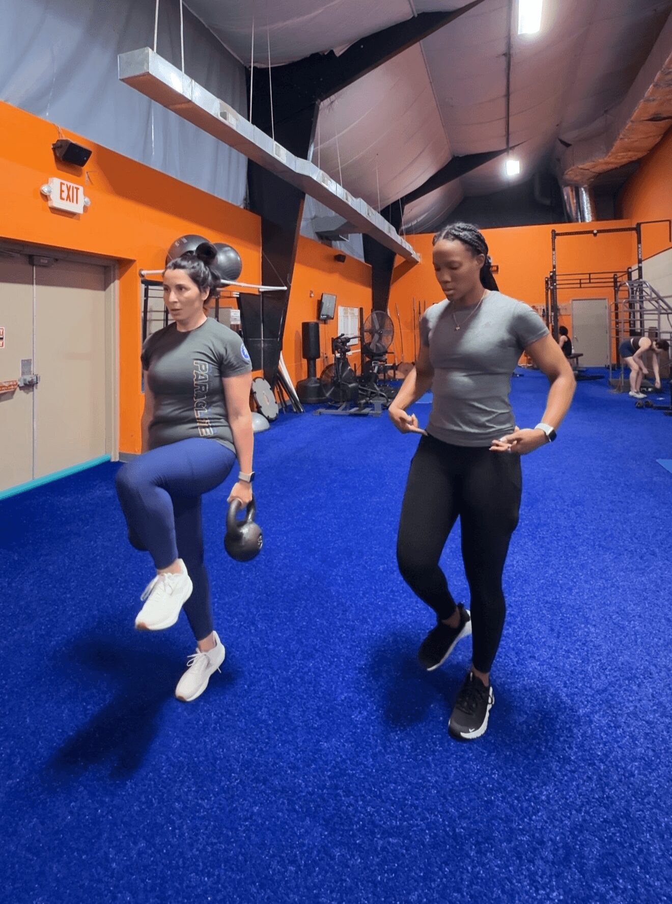 Two women exercising in a gym with blue flooring and orange walls, one lifting a kettlebell, the other walking.