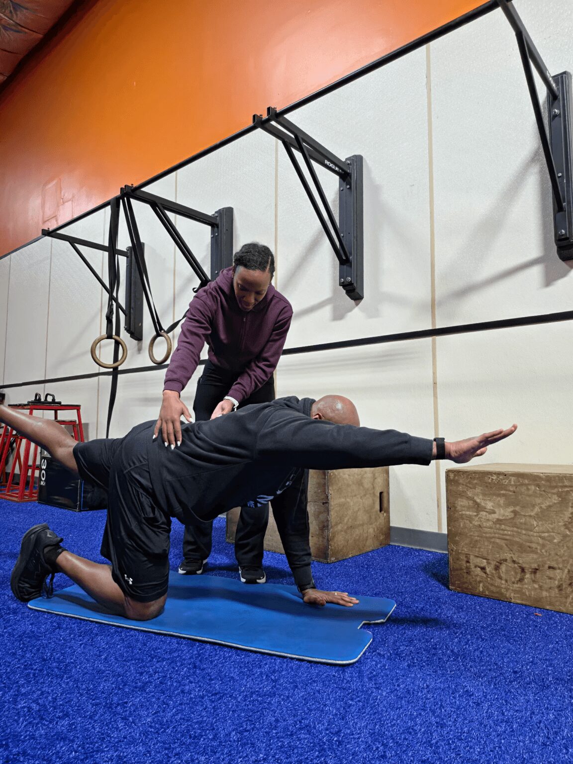 Person in black leggings and a purple top performs a yoga pose on a blue mat, with a trainer assisting in a gym.