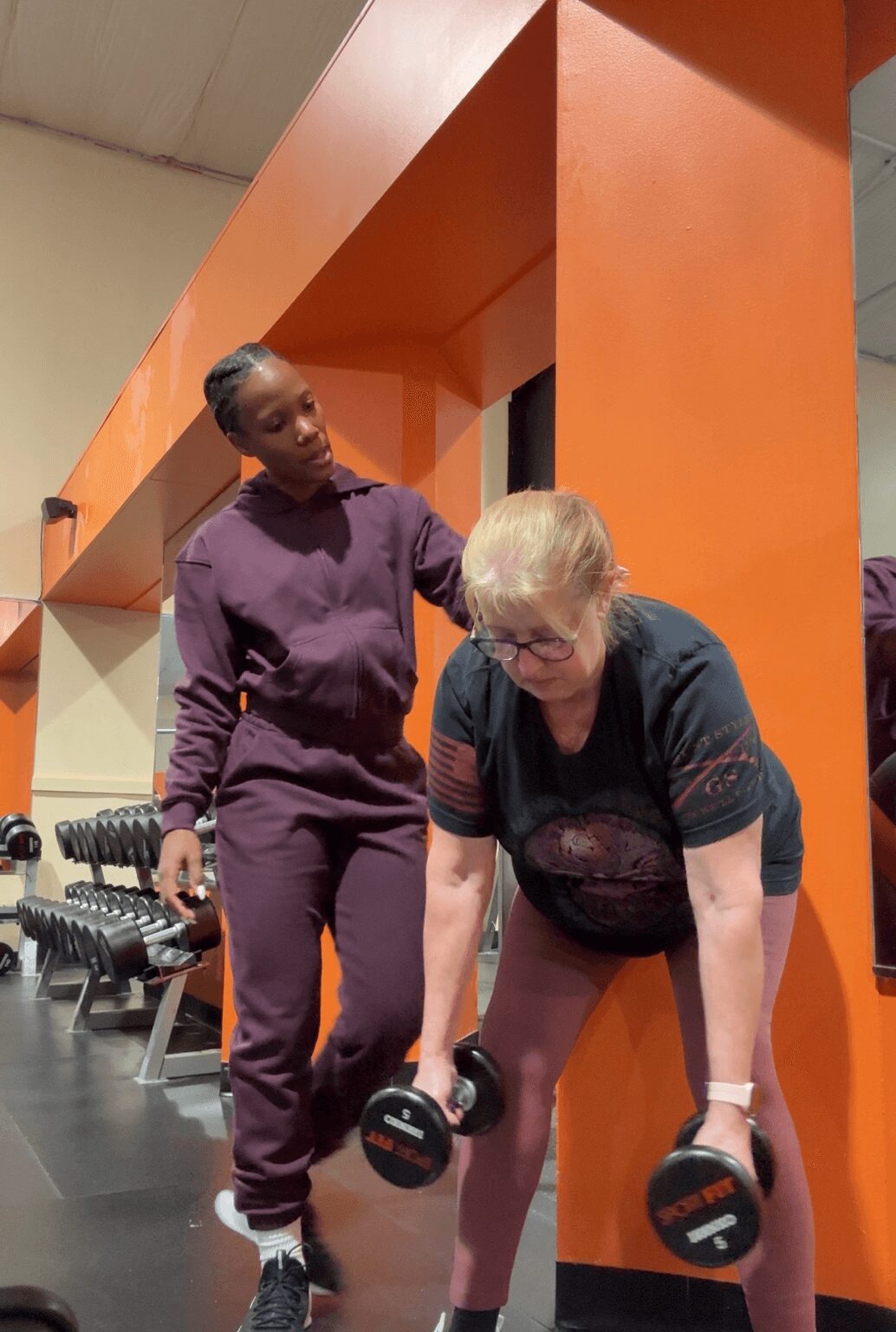 A woman with glasses and a black shirt is lifting dumbbells while a trainer in purple observes in a gym.