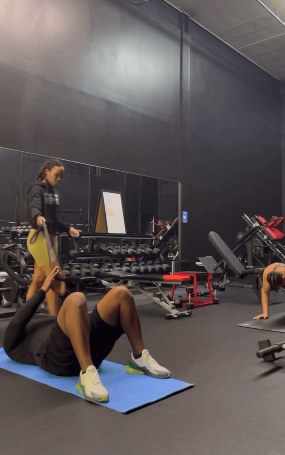Person lying on a blue exercise mat in a gym, with another person standing nearby, gym equipment in background.