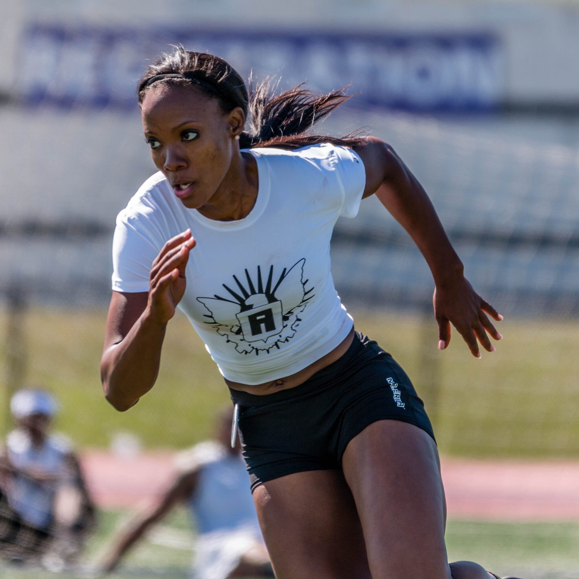 Female athlete running on track, wearing white t-shirt and black shorts, outdoors during daytime.