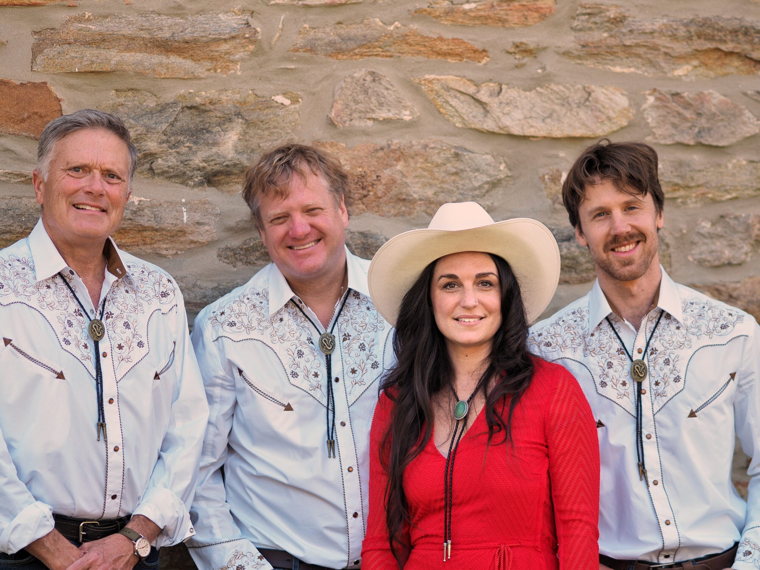 Four people standing outdoors in front of a stone wall, smiling, with one woman wearing a white hat and red jacket.