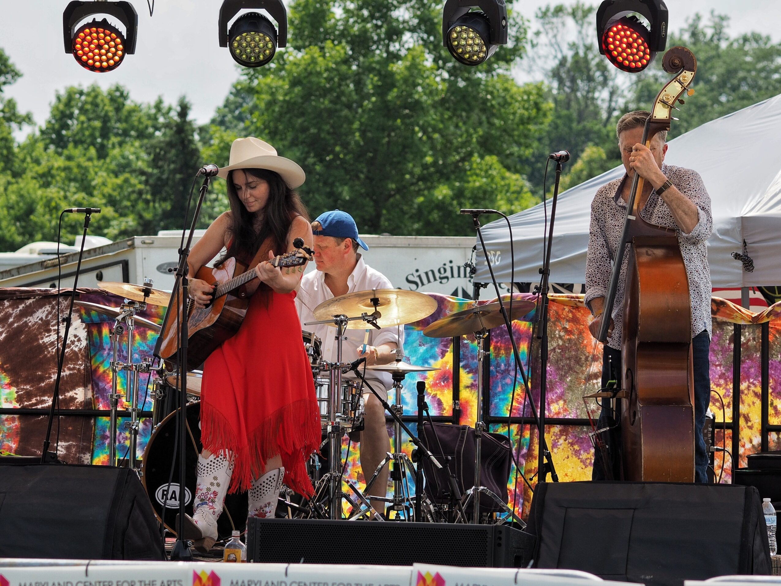 Three musicians perform on an outdoor stage with trees and tents in the background, including a woman with a guitar, a drummer, and a man with a double bass.