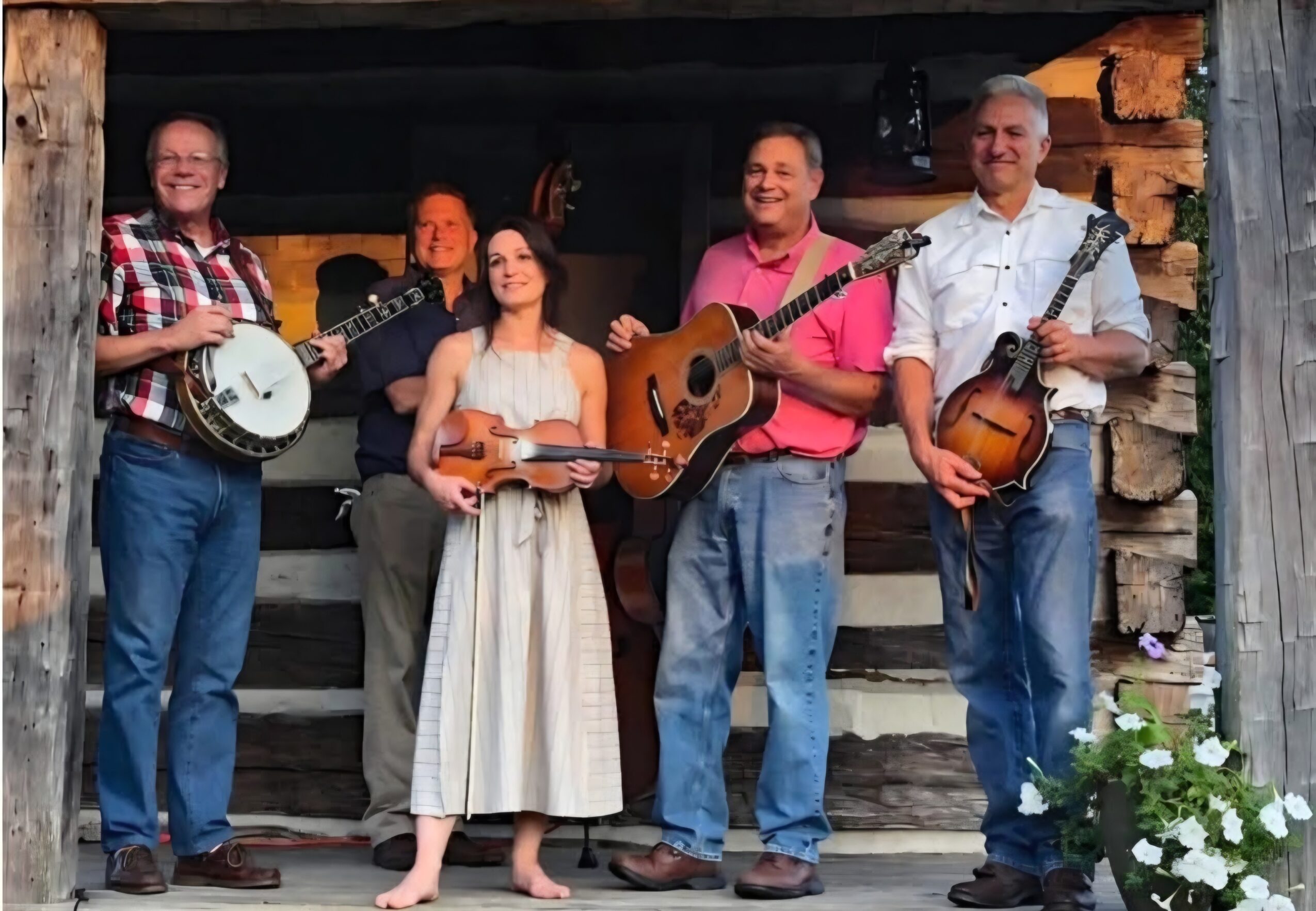Group of six people with musical instruments standing outdoors in front of a log cabin, smiling.