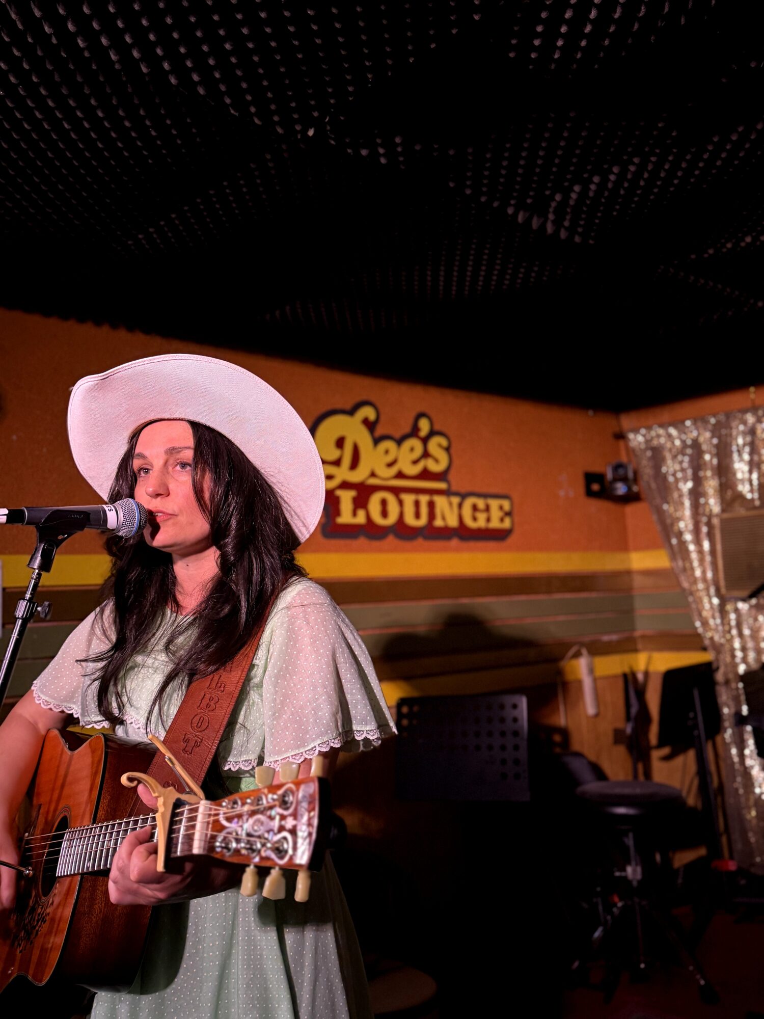 Woman with long dark hair wearing a white hat and gray dress, playing guitar and singing at a microphone in a lounge.