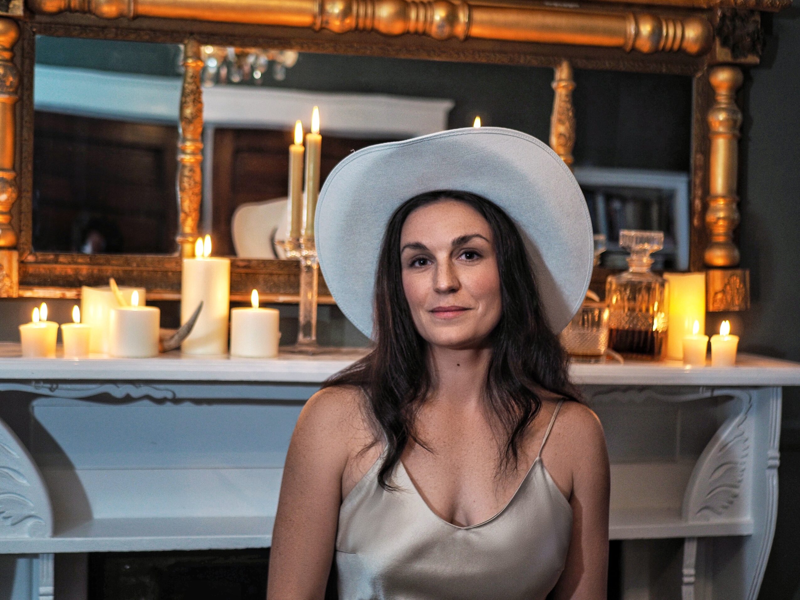Woman wearing a large white hat and satin dress, standing in front of a decorated fireplace with candles.