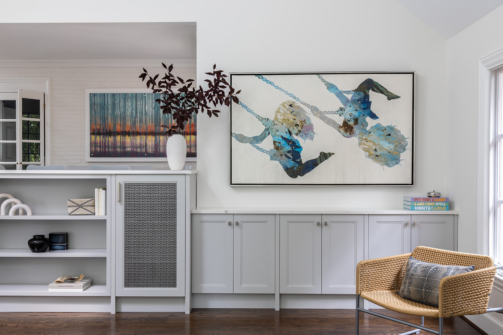 Living room with artwork, a cabinet, a wicker chair, and a window, decorated with a vase and books.