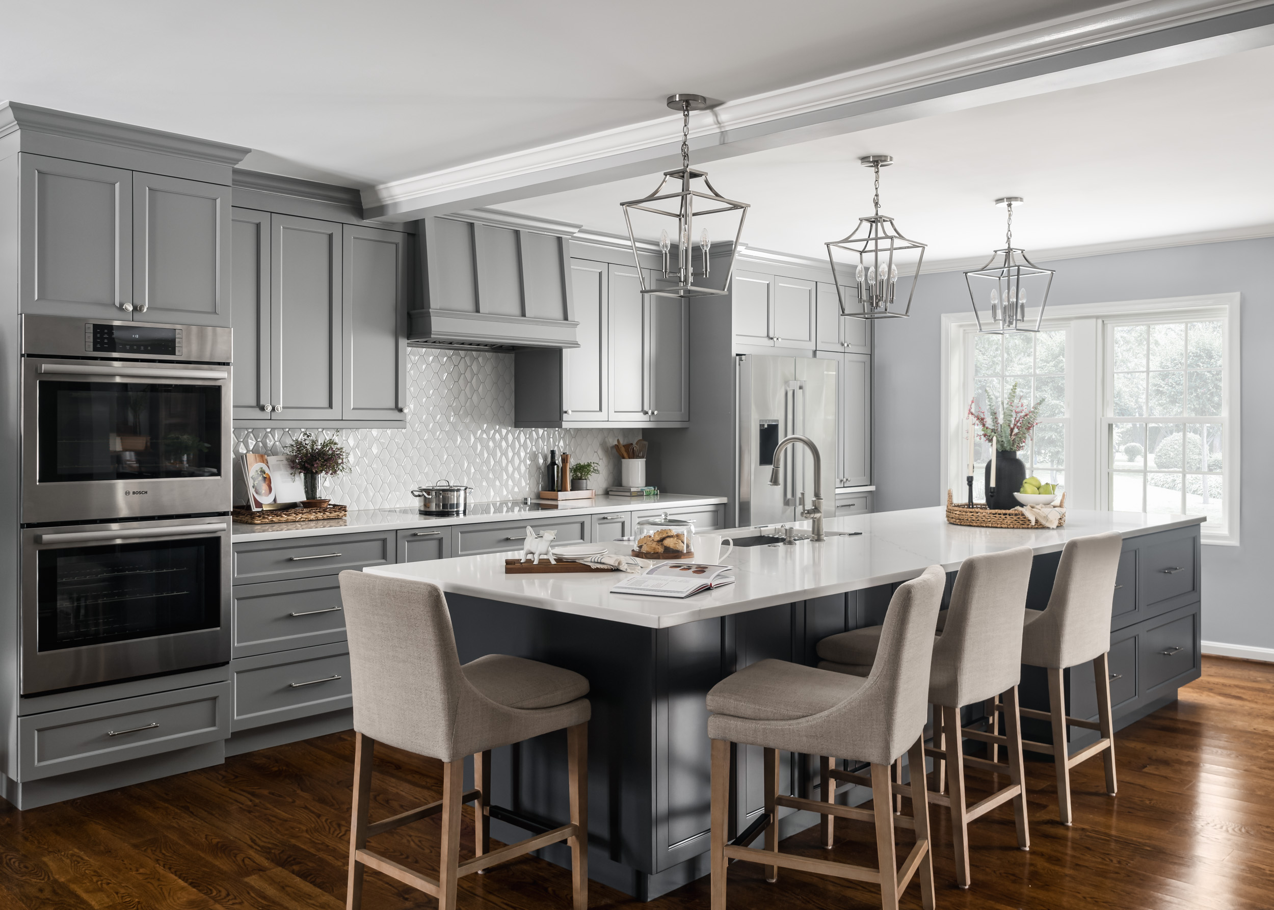 Modern kitchen with gray cabinets, white island, and four beige chairs, bright with large windows and pendant lights.