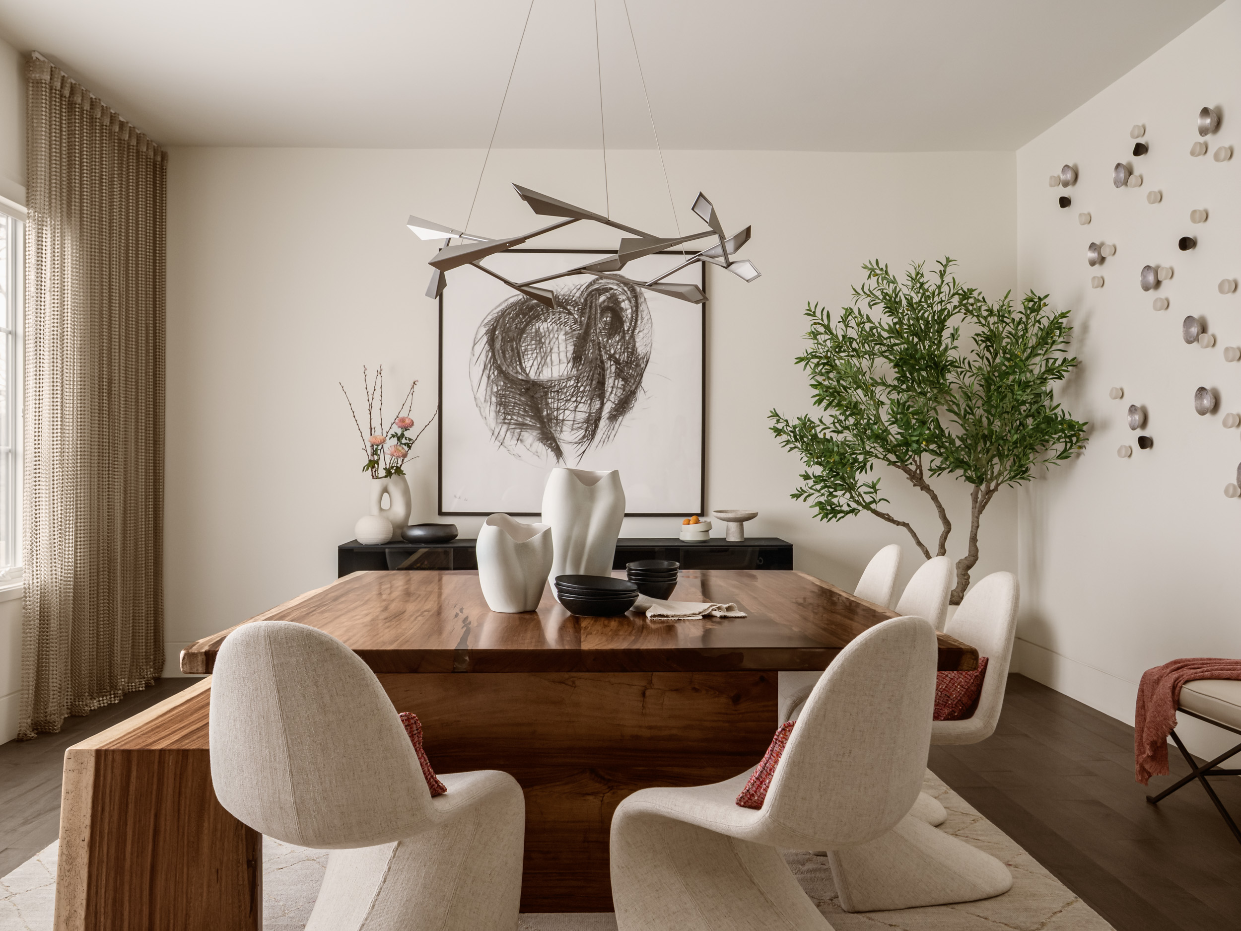 Dining room with wooden table, six chairs, a large plant, and wall art, illuminated by natural light from window.