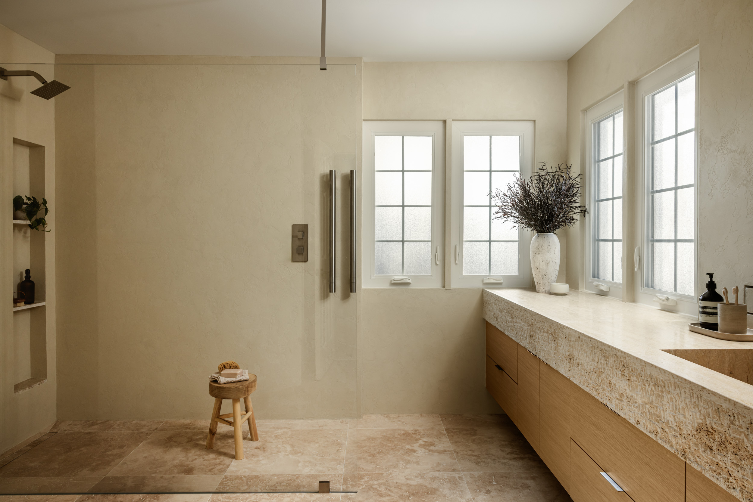 Bathroom with windows, a vase with flowers, a countertop, and a small stool, in neutral tones.