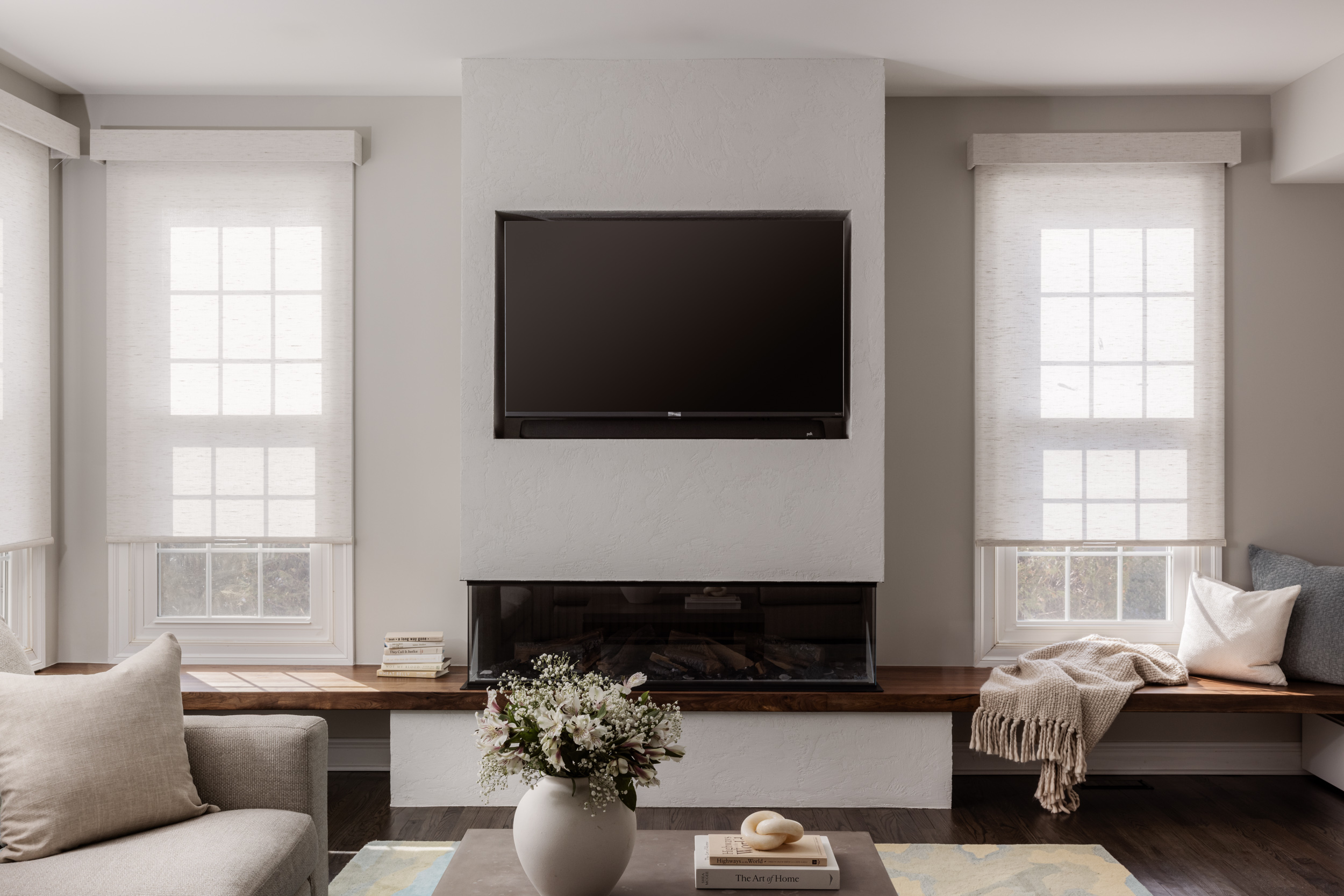 Living room with two large windows, a wall-mounted TV, a sofa, and a window seat with cushions and a blanket.