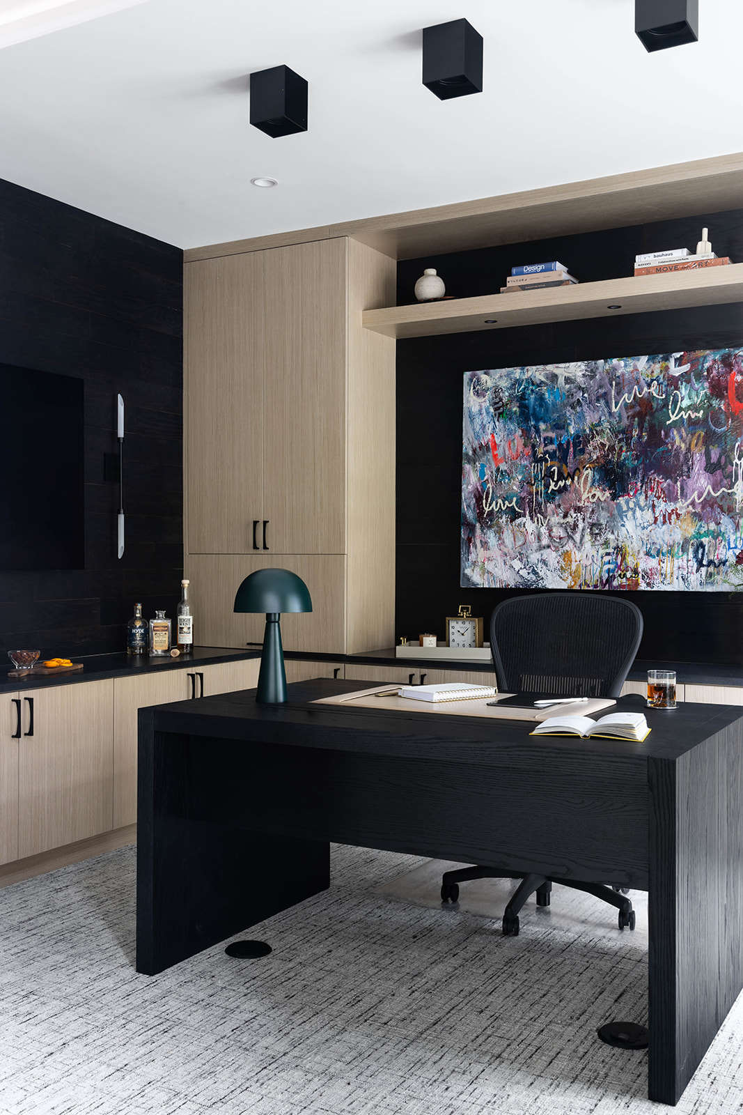 Modern office with black desk, chair, and wall, light wood cabinets, and abstract artwork on screen, ceiling lights, and shelves.
