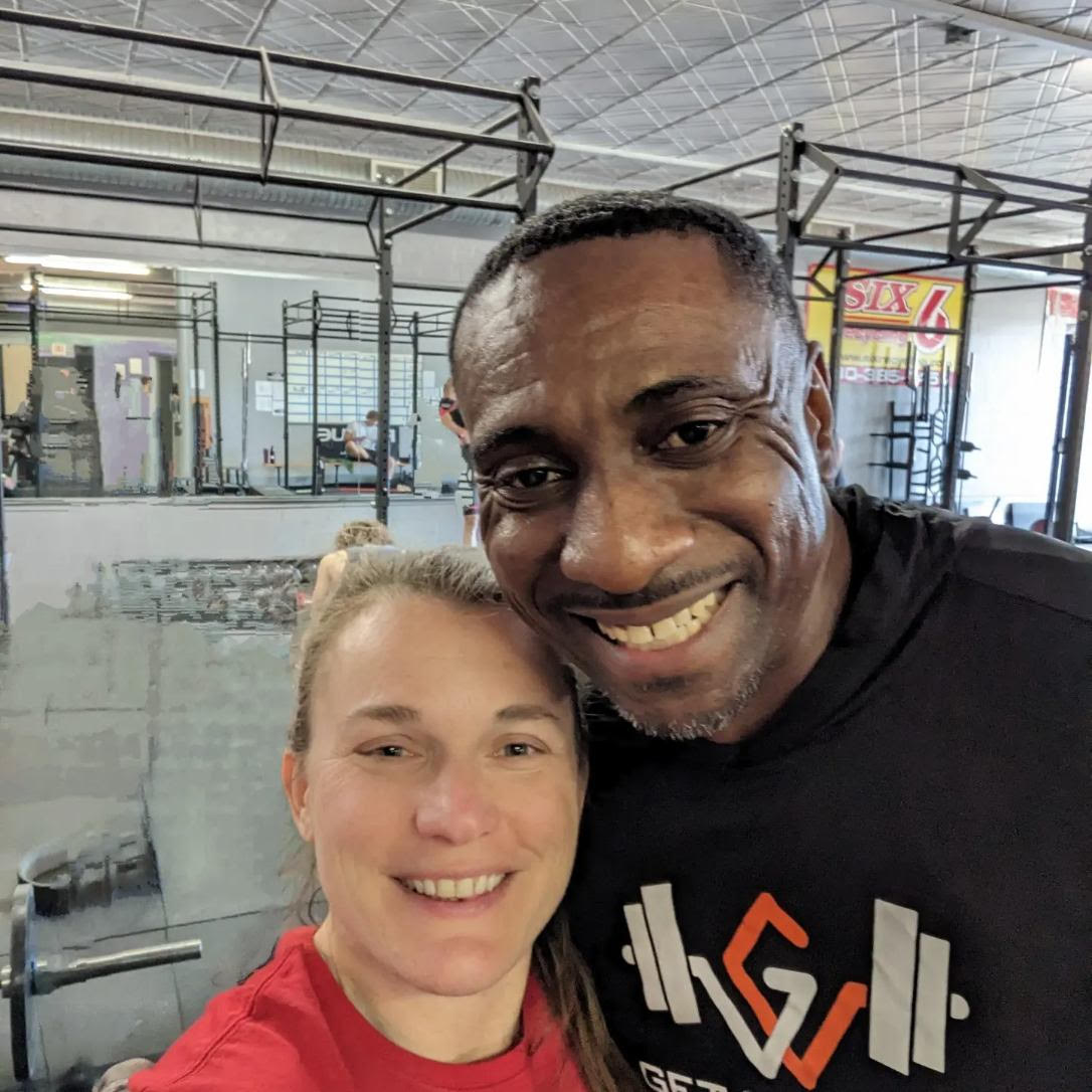 Two people smiling indoors, one with blonde hair and the other with short dark hair, in a gym or fitness center.