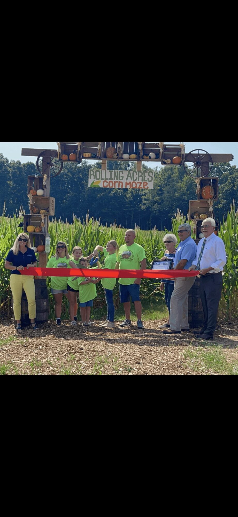 Group of people at a ribbon-cutting event outdoors, with a sign reading 'Growing Acres' and trees in the background.