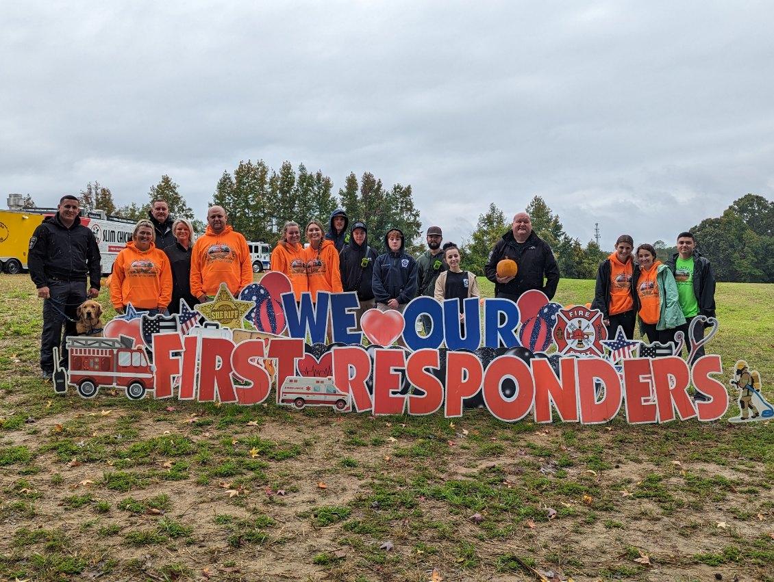 Group of people holding a colorful sign that reads 'We Love Our First Responders' on a grassy field with trees and cloudy sky in background.