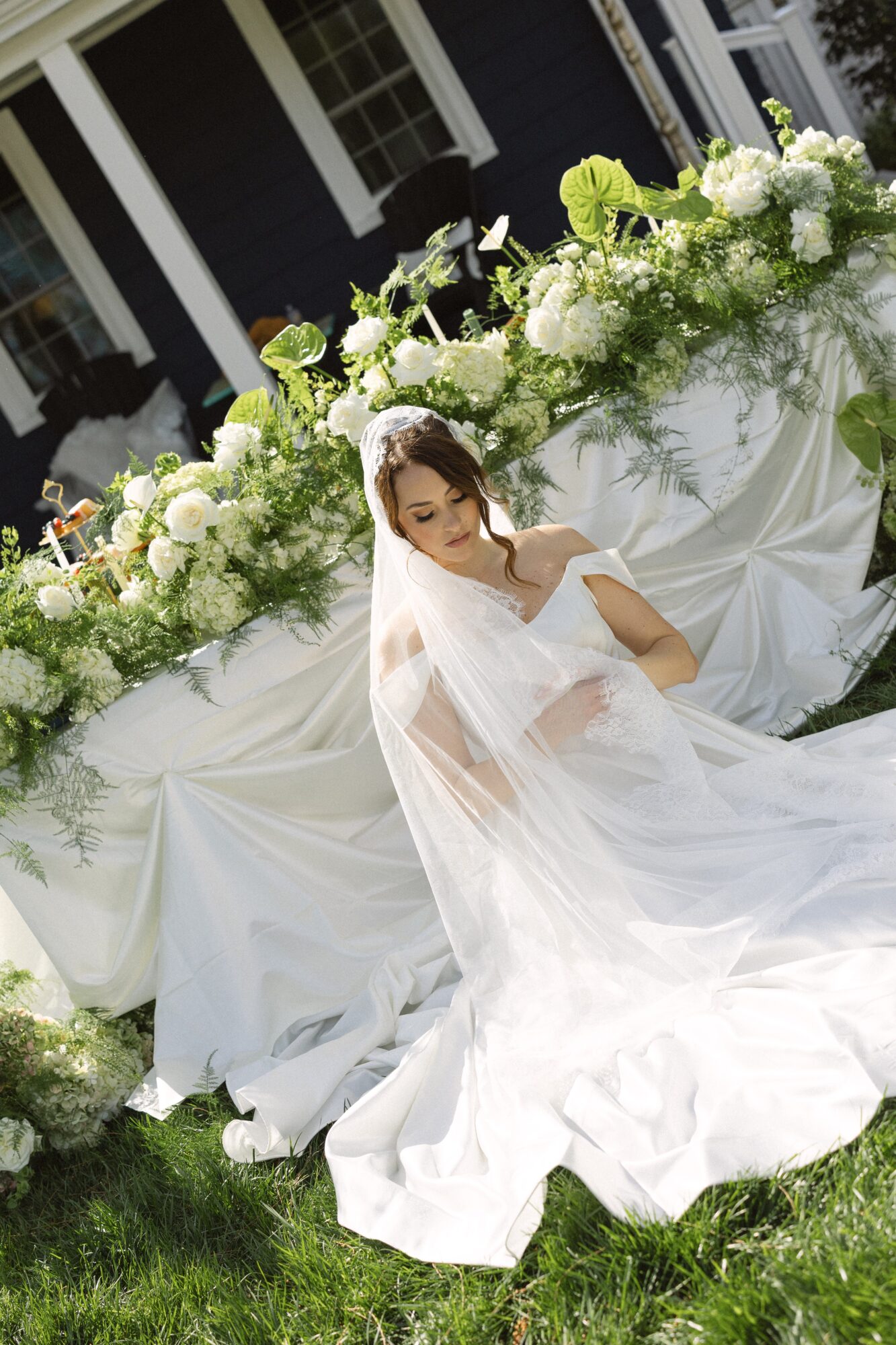 Bride in white dress and veil sitting outdoors near floral arrangement with white and green flowers, on grass.