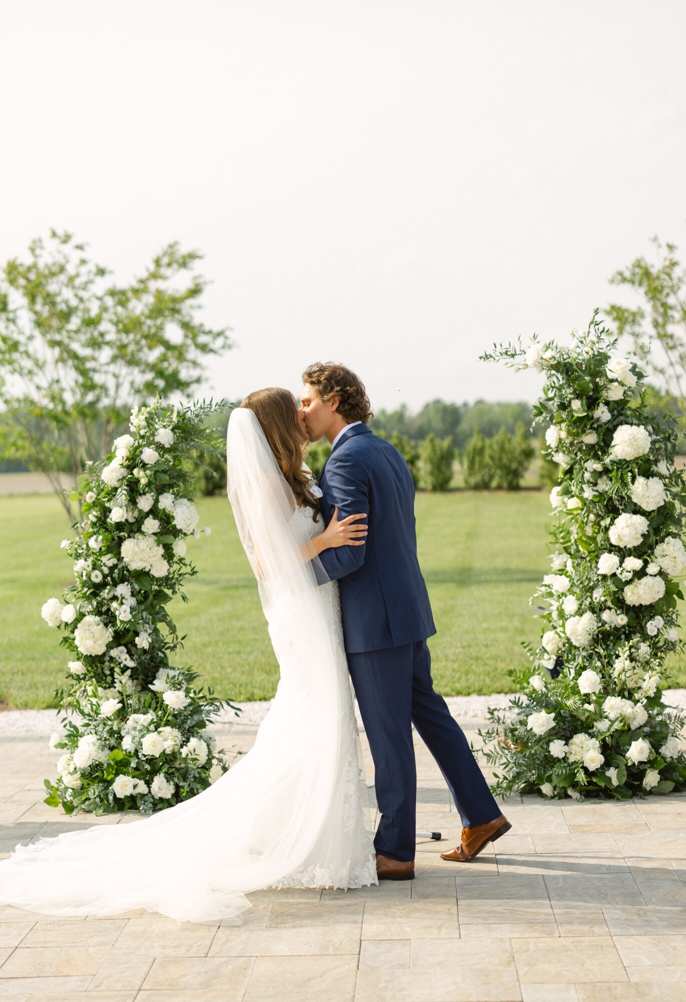 Bride and groom kiss outdoors with floral arches, green trees, and a grassy field in the background.