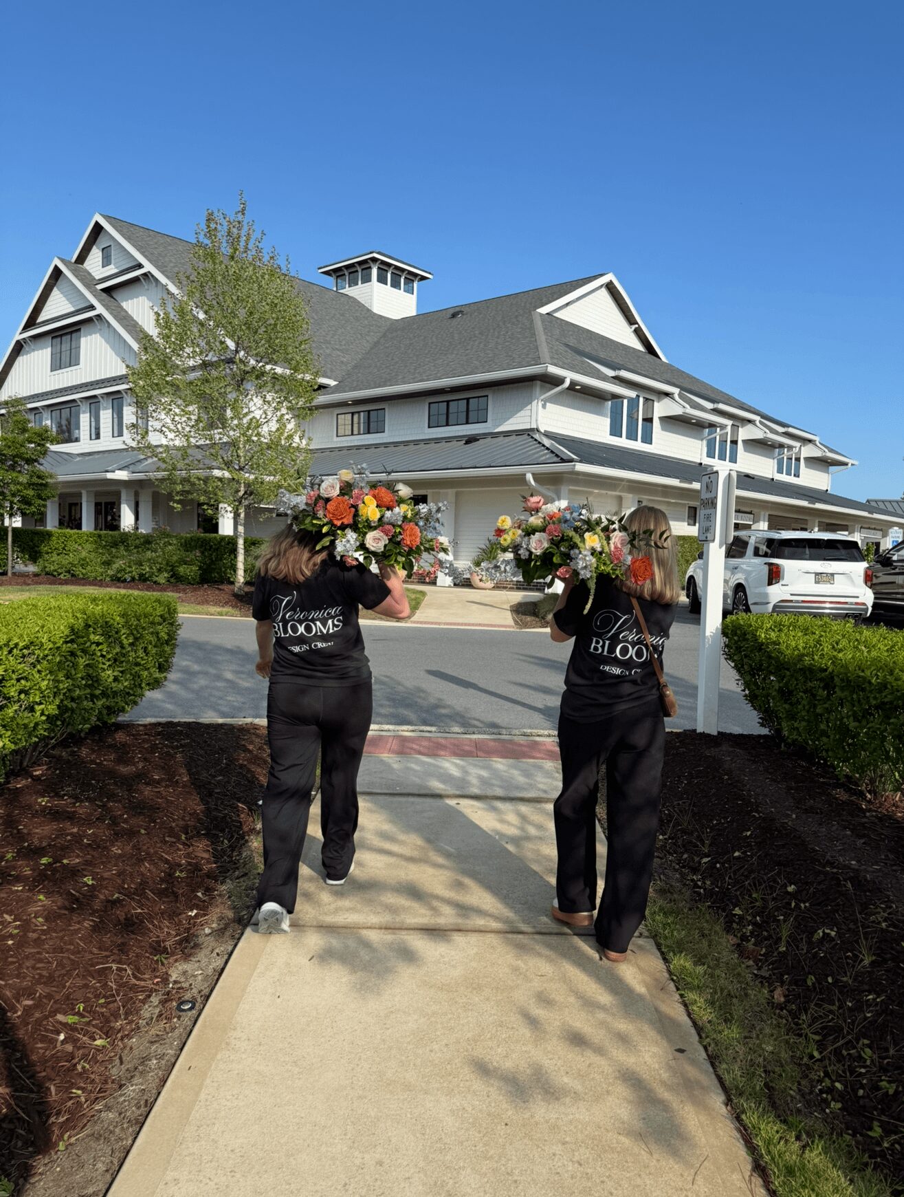 Two people carrying large flower arrangements walk on a sidewalk toward a large white building with multiple gables and a tower, under a clear blue sky.