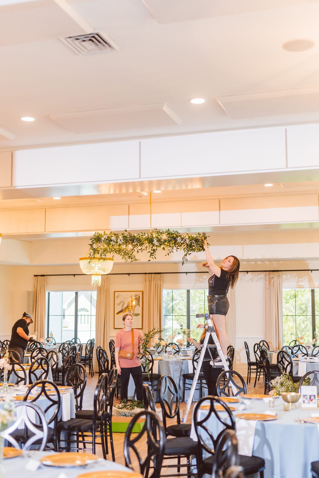 People setting up decorations in a spacious, well-lit banquet hall with round tables and chairs, large windows, and hanging greenery.