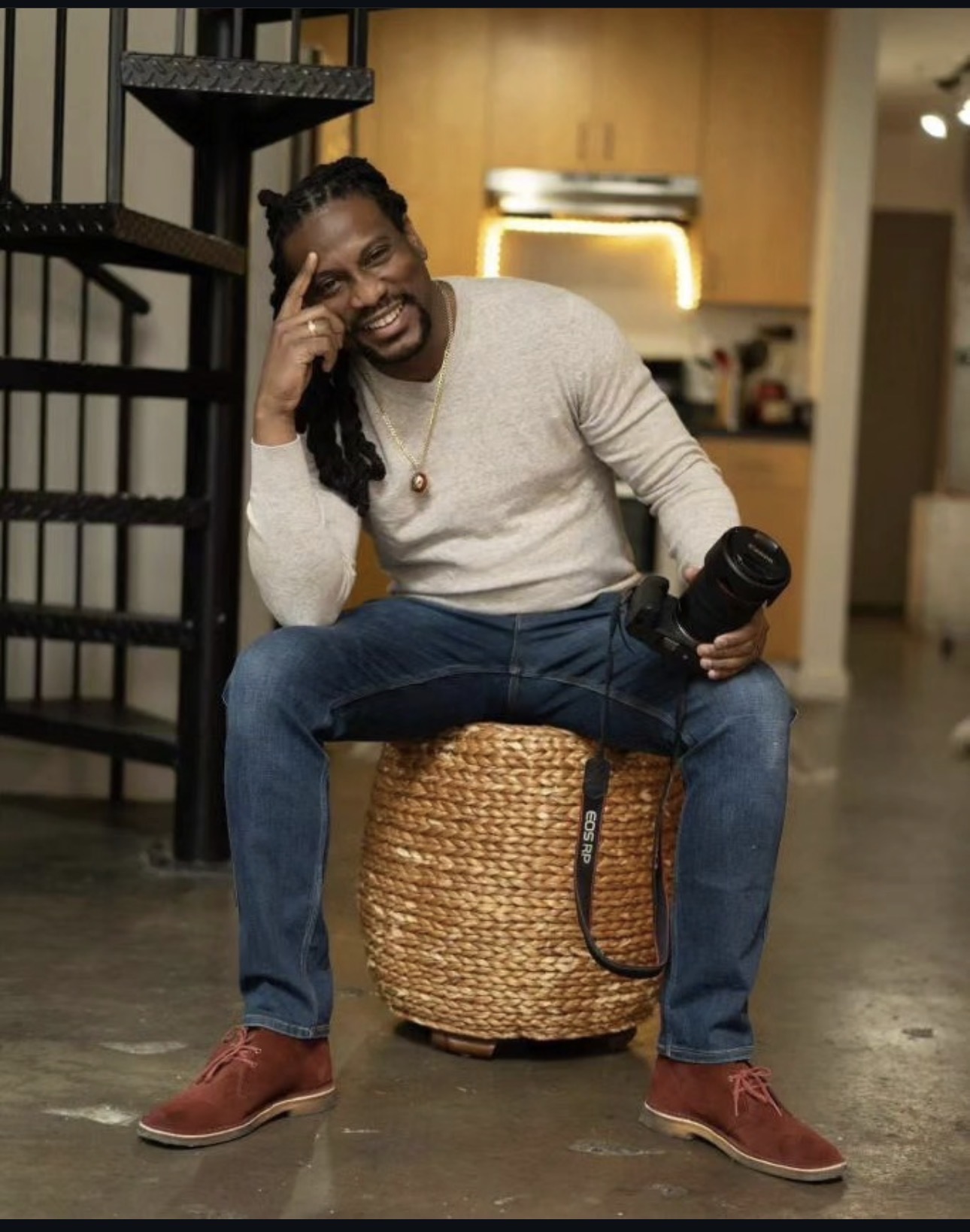 Person sitting on a woven basket, smiling, holding a camera, in a home interior with a staircase and kitchen in background.
