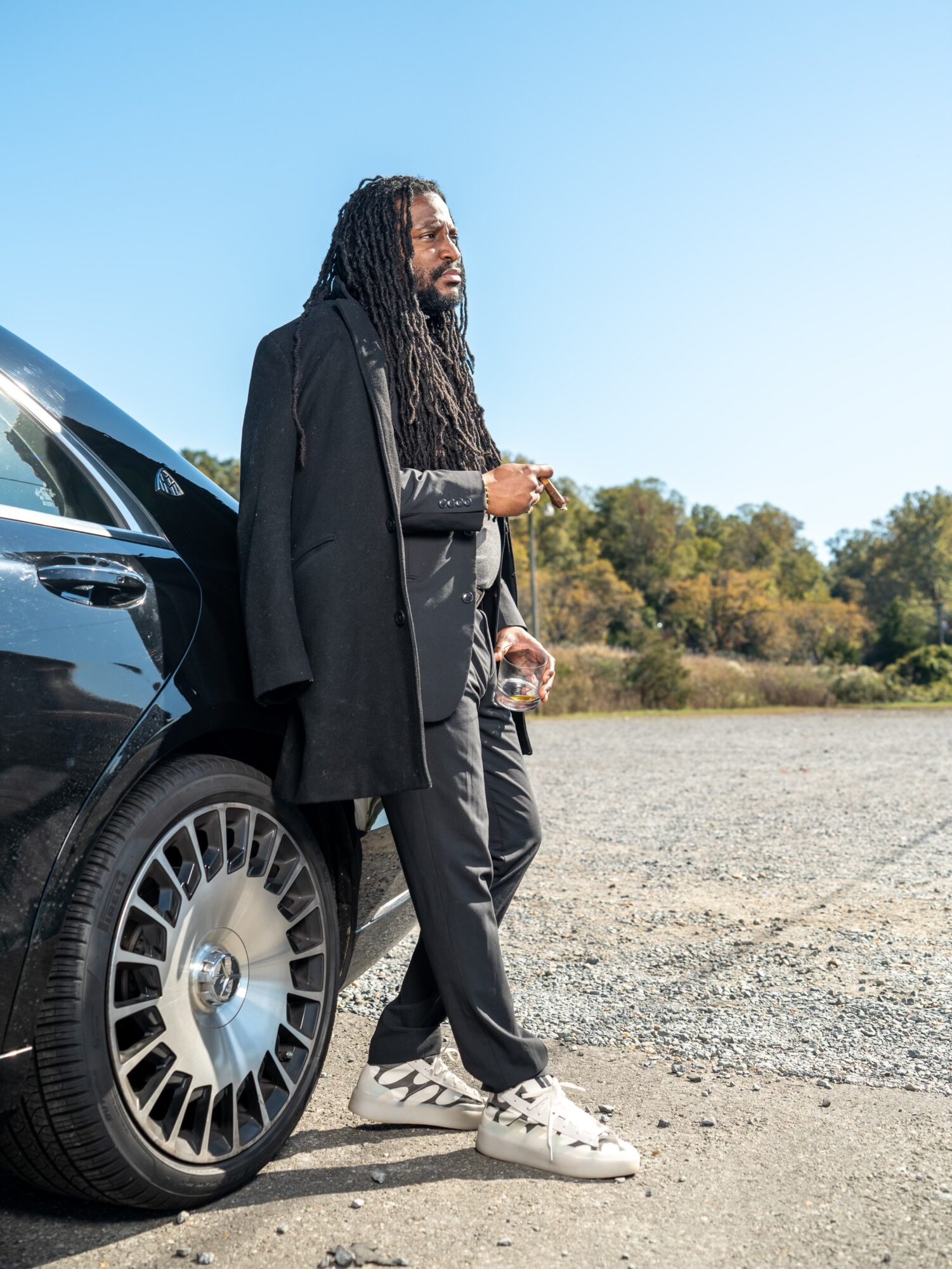 Man with long hair and beard leaning against a black car outdoors, wearing a dark coat and gray pants, with trees in the background.