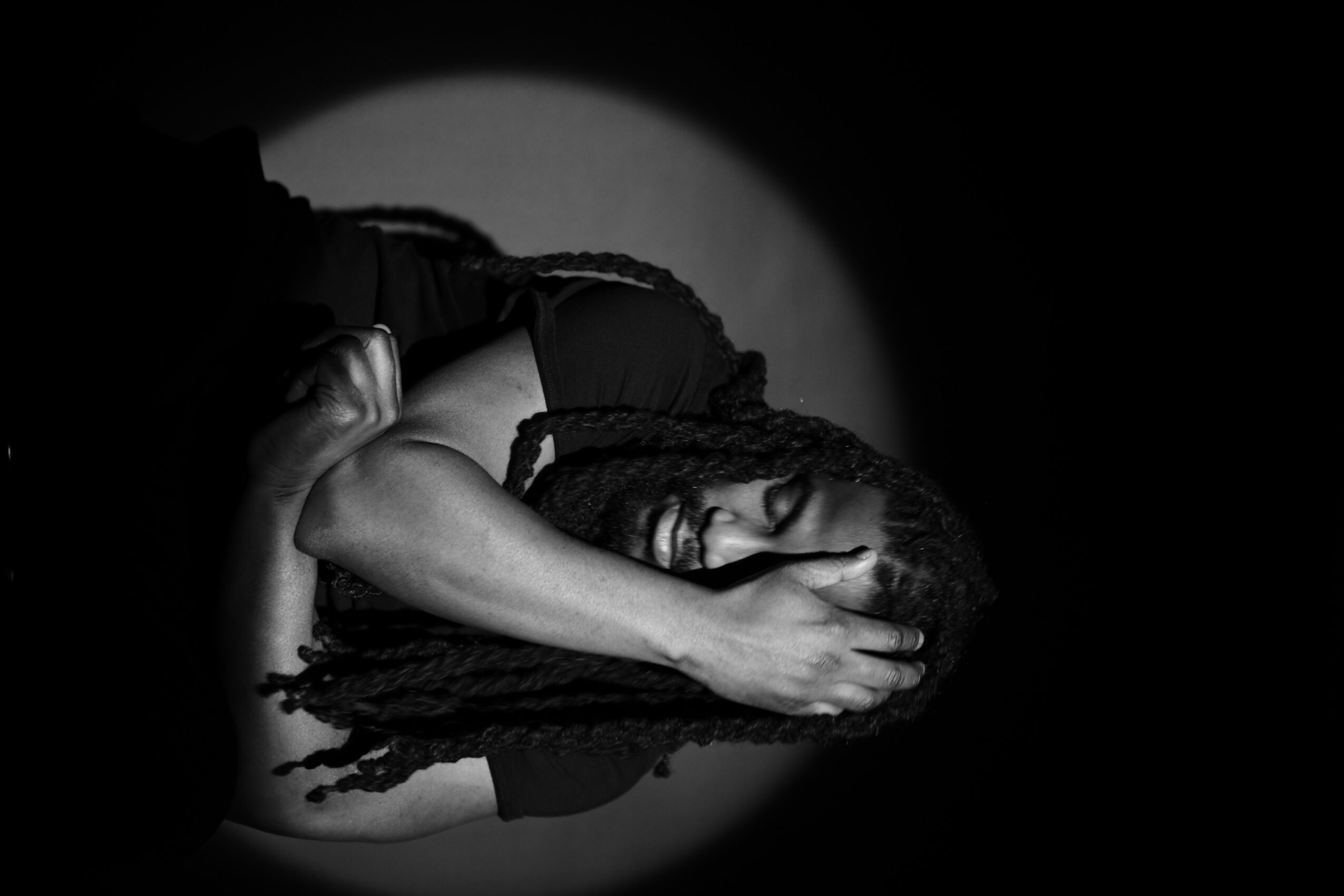 Person with long dreadlocks covering face, lying down with hand on head, in black and white, spotlight effect