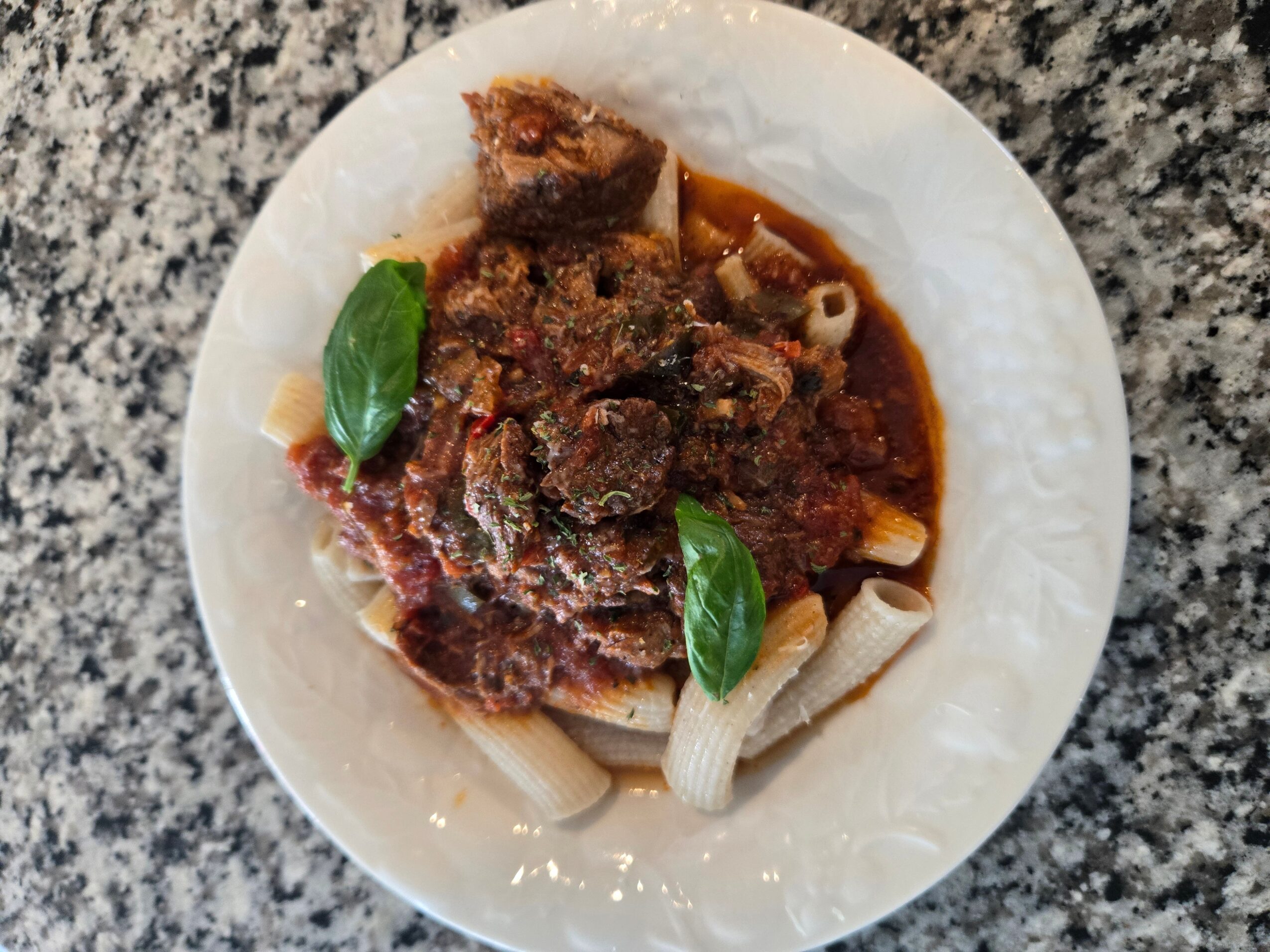 Plate with cooked meat, pasta, and basil leaves on a granite countertop.