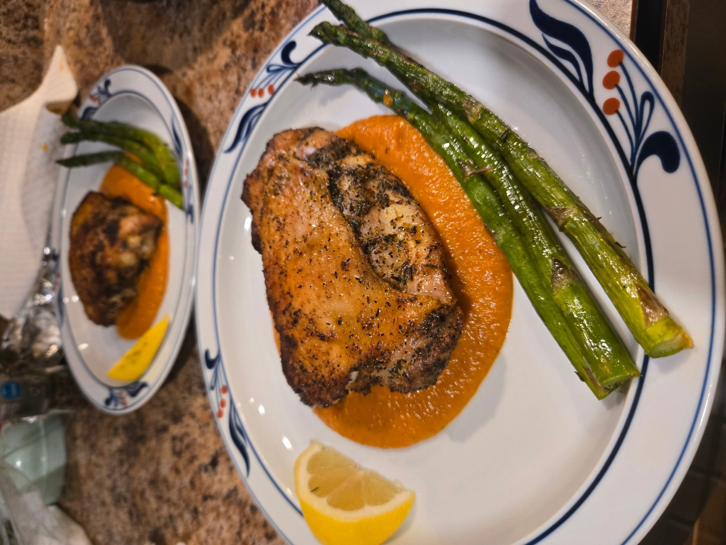 Grilled chicken thigh with orange sauce, served with asparagus and lemon wedge on a decorative plate.