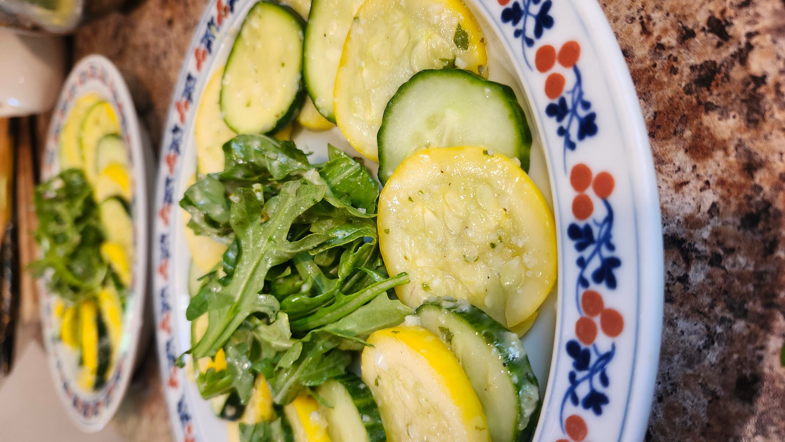 Plate with sliced cucumbers, yellow squash, and leafy greens on a patterned dish, on a brown surface.