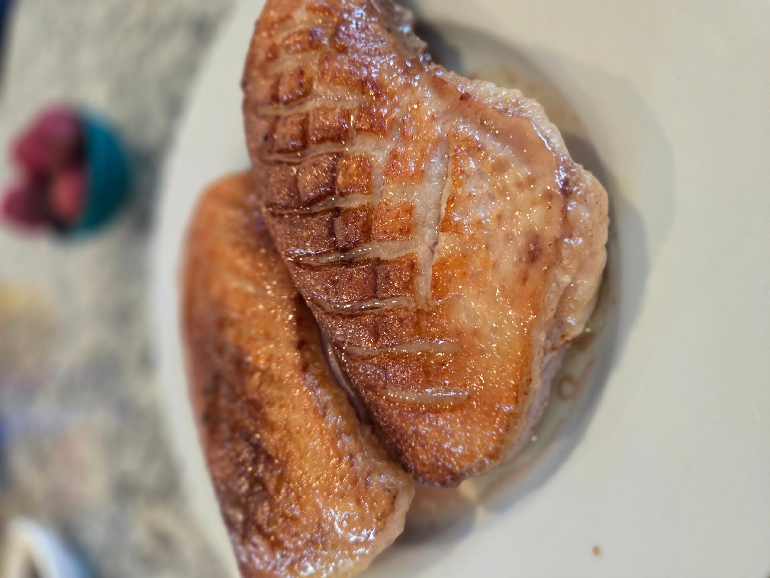 Two cooked chicken drumsticks on a white plate, with a blurred background including a small container of raspberries.