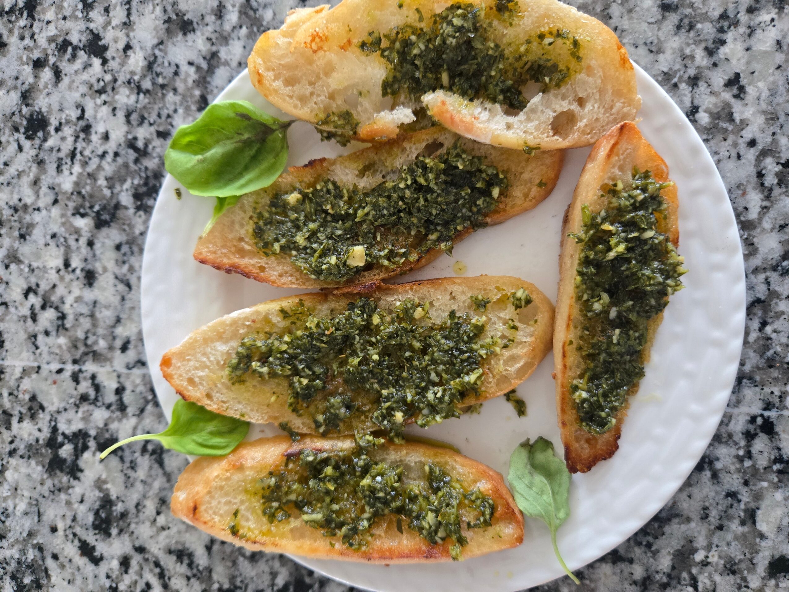 Six slices of toasted bread topped with green herb spread, garnished with basil leaves, on a white plate on a granite surface.