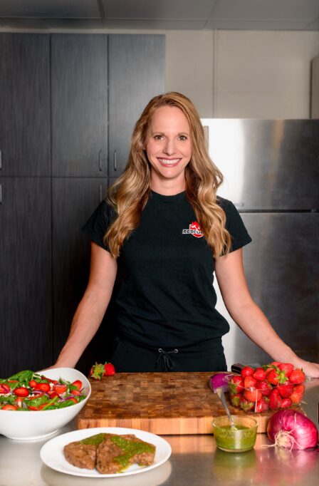 Young woman with long wavy hair smiling, standing behind a wooden cutting board with vegetables and a plate of food.