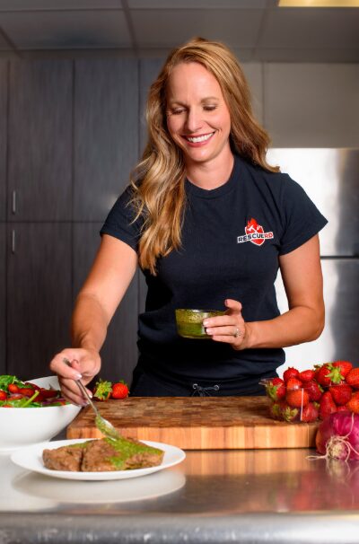 Woman with long wavy hair smiling, preparing food in a kitchen, holding a small bowl, surrounded by vegetables and a plate of food.