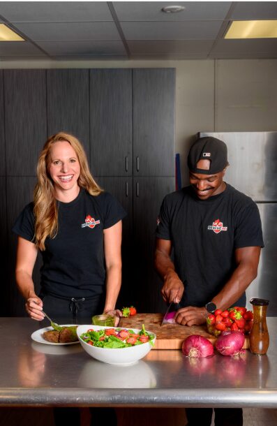 Two people preparing food in a kitchen, with a bowl of salad and vegetables on the counter.